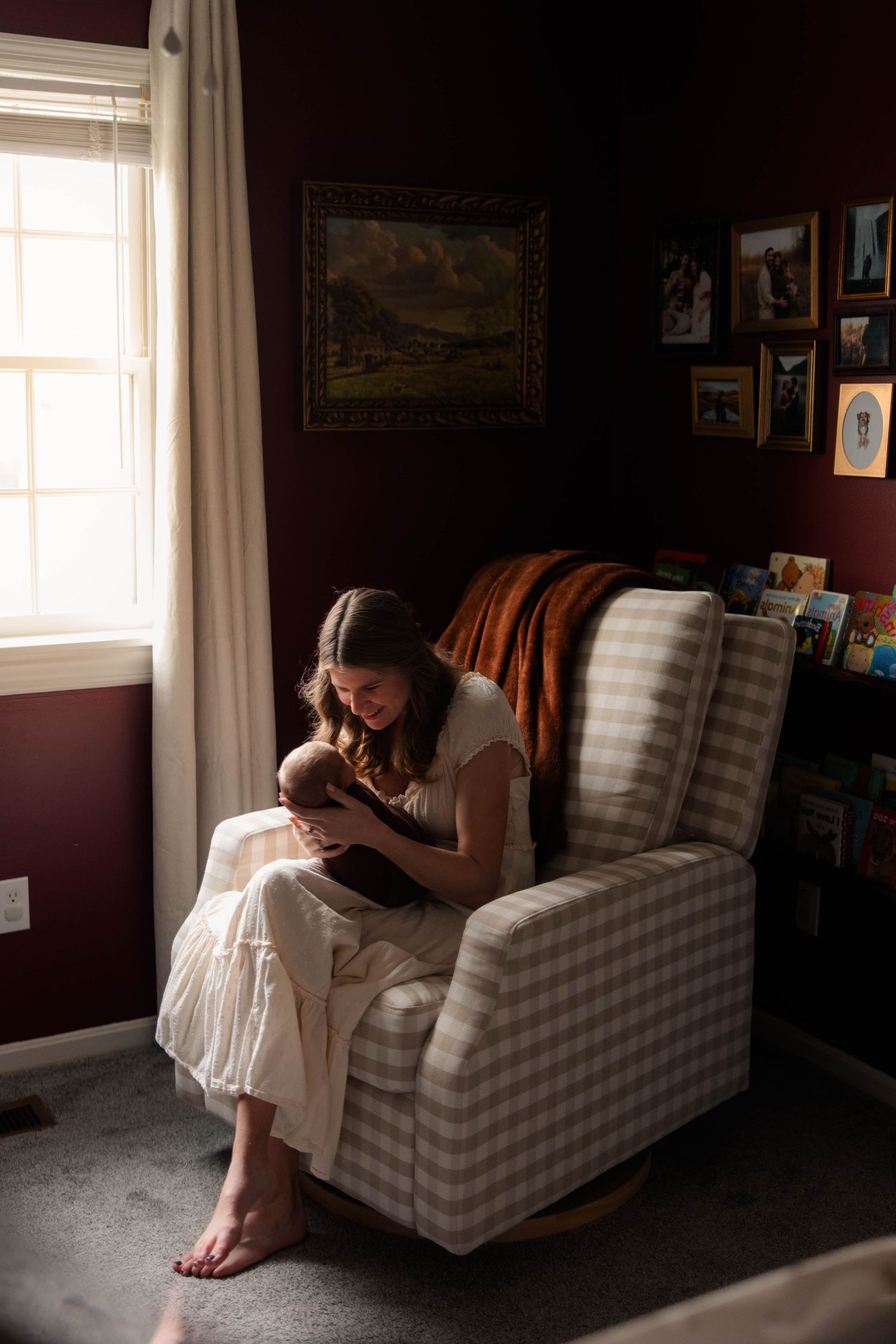 Mother sitting in a plaid glider chair cradling her newborn baby by a sunlit window in a nursery.