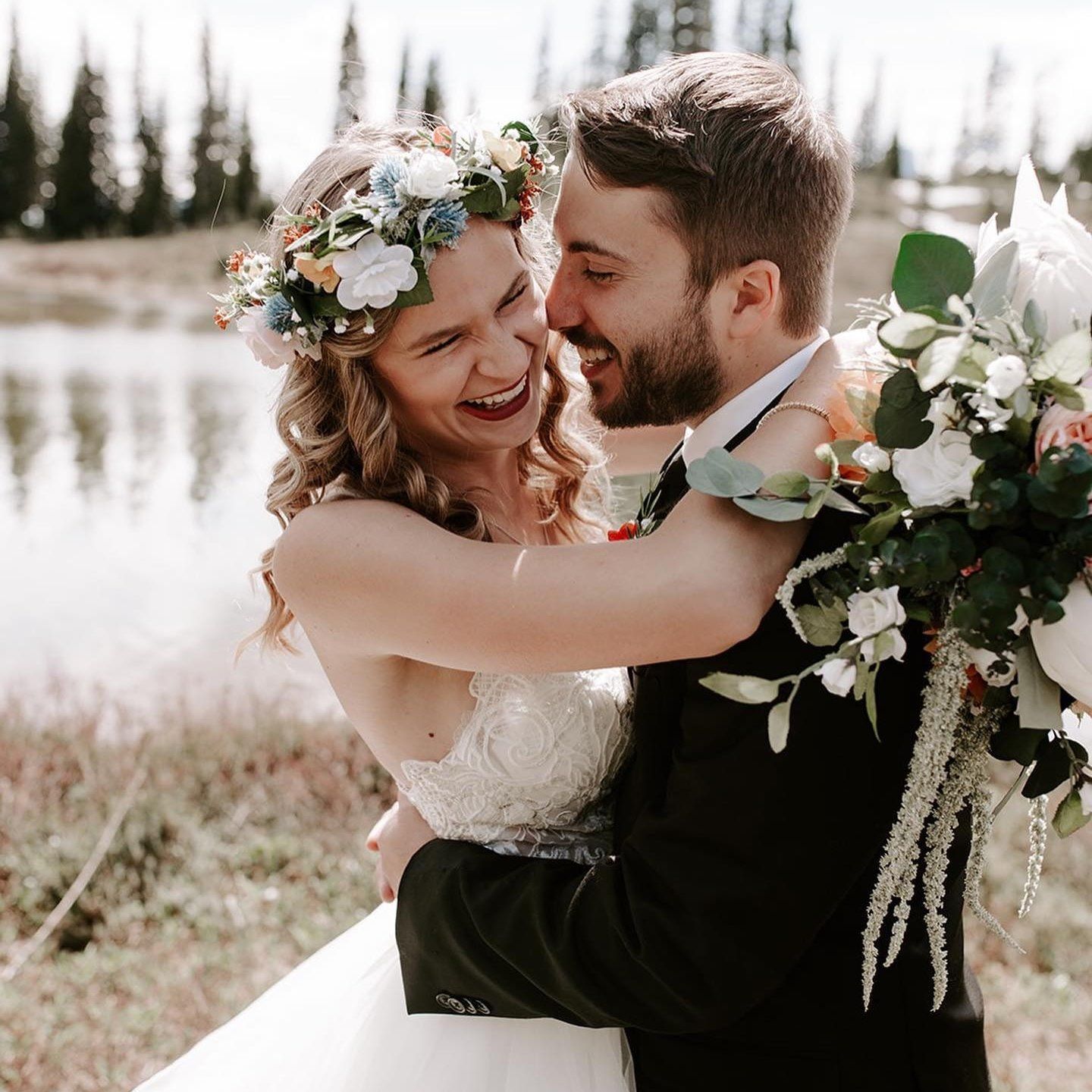 Joyful bride with floral crown laughing as groom lifts her during outdoor wedding celebration.
