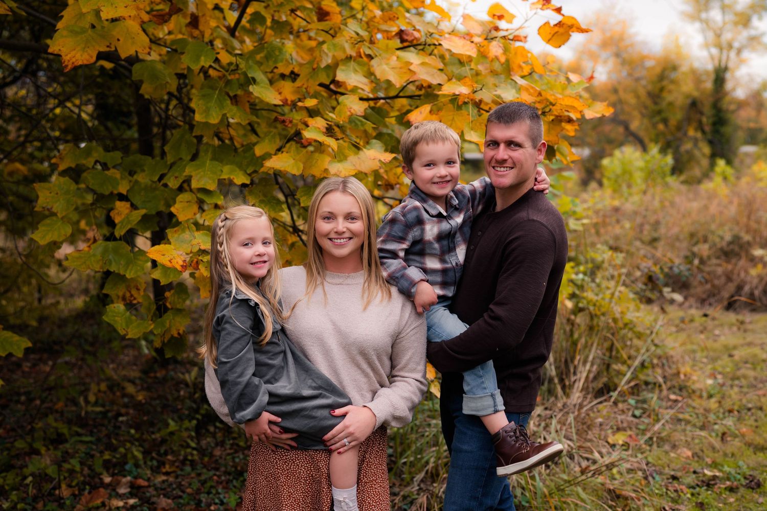 Family of four smiling outdoors in autumn, parents holding children under colorful fall foliage.