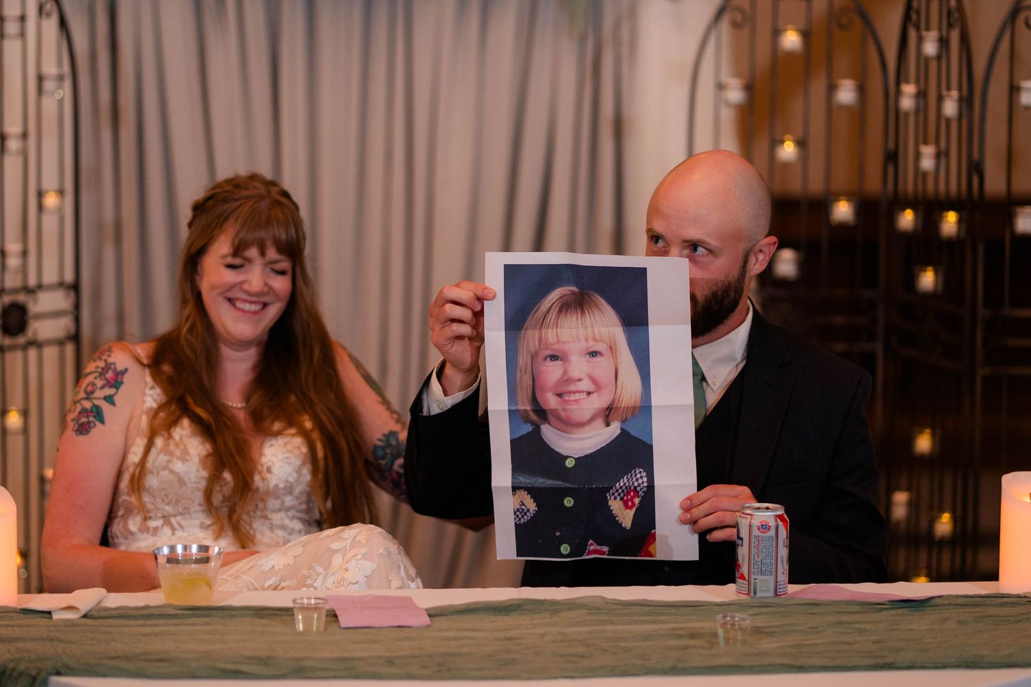 Bride and groom laughing as groom holds up a childhood photo during wedding reception speeches.