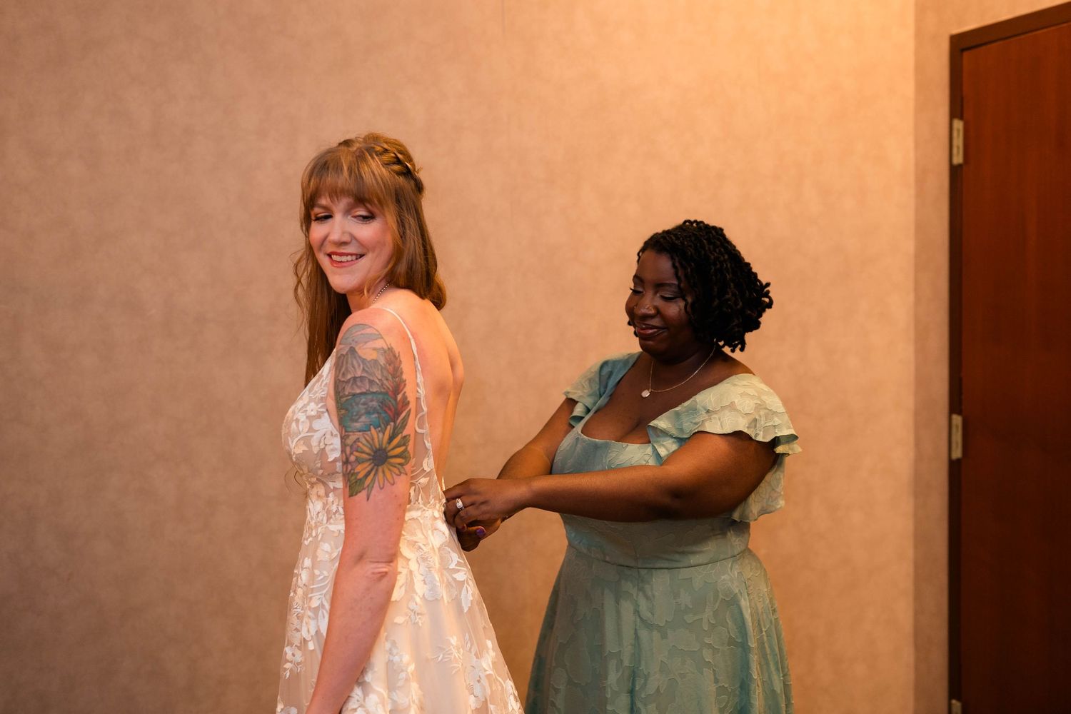 Bride in white lace wedding gown being helped by bridesmaid in sage green dress.