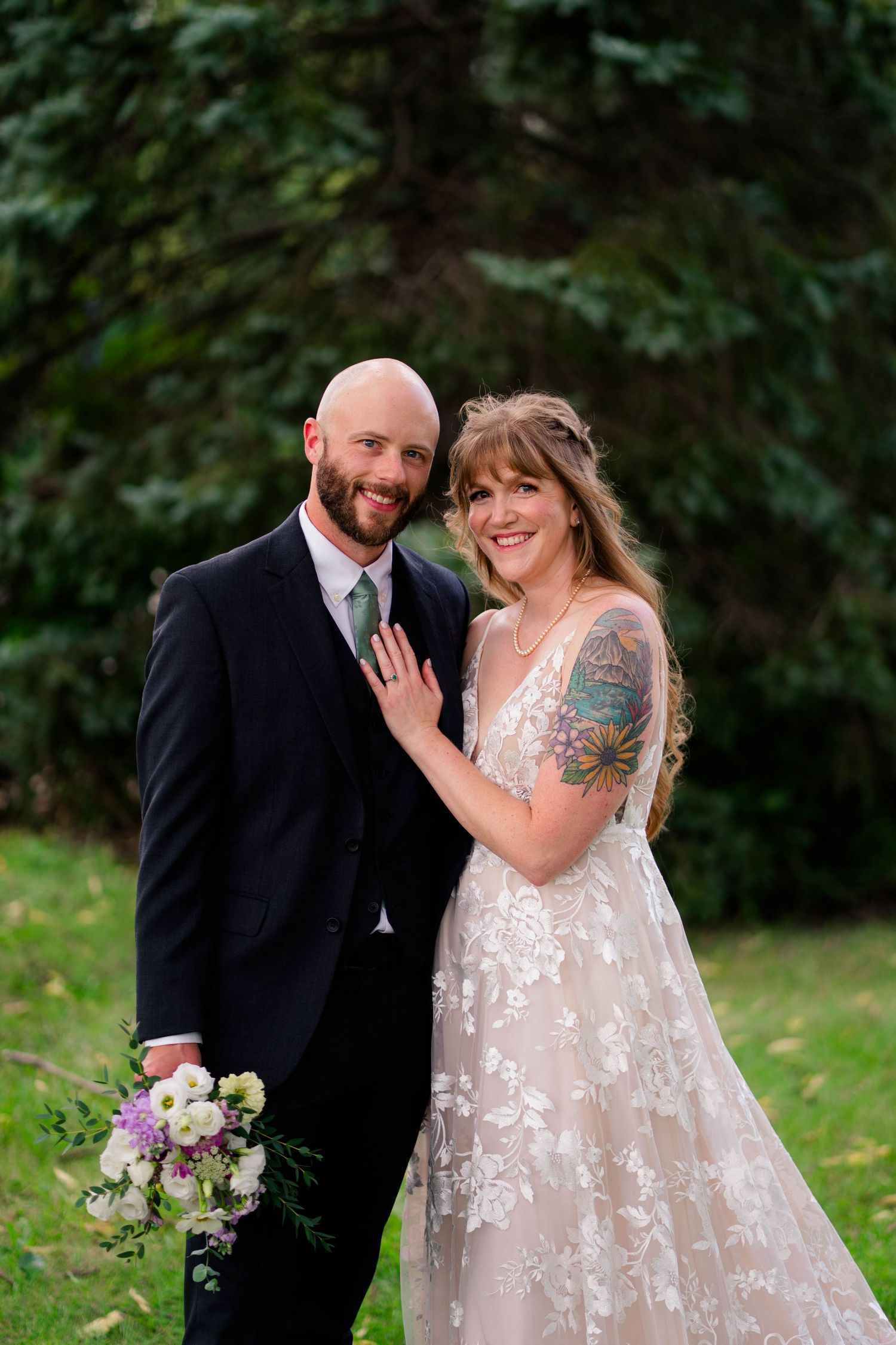 Happy bride and bald groom posing together outdoors, bride in lace gown with floral tattoo holding bouquet.