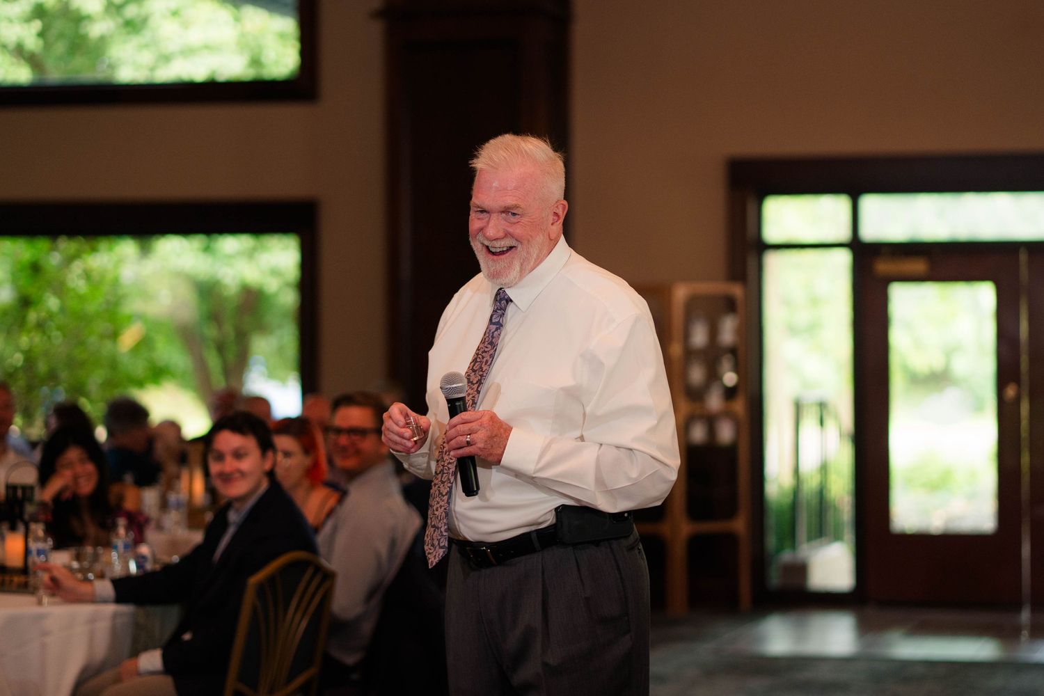 Older man smiling while giving toast with microphone at elegant wedding reception venue.