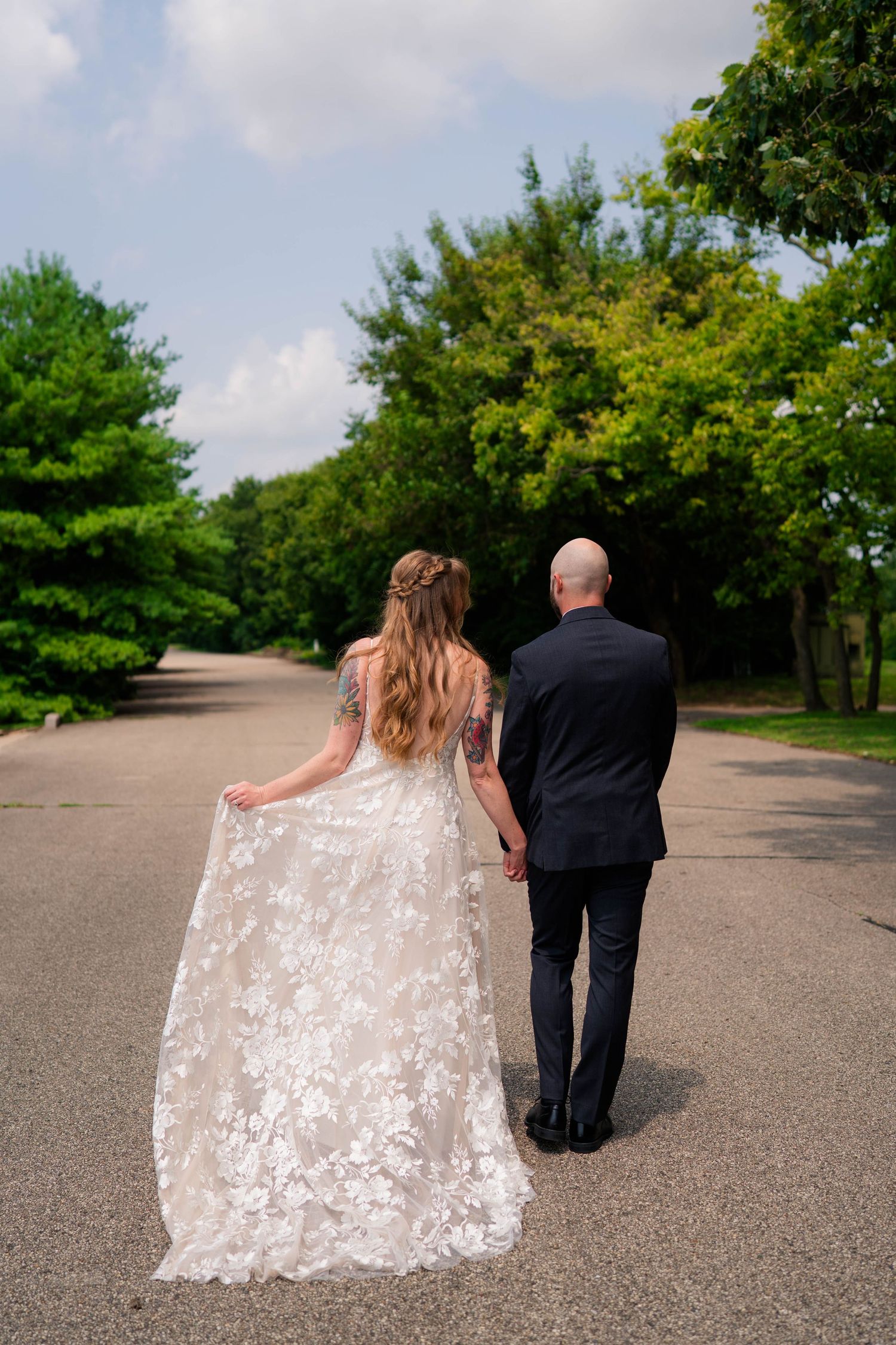 Bride and groom walk hand in hand down tree-lined path in wedding attire.