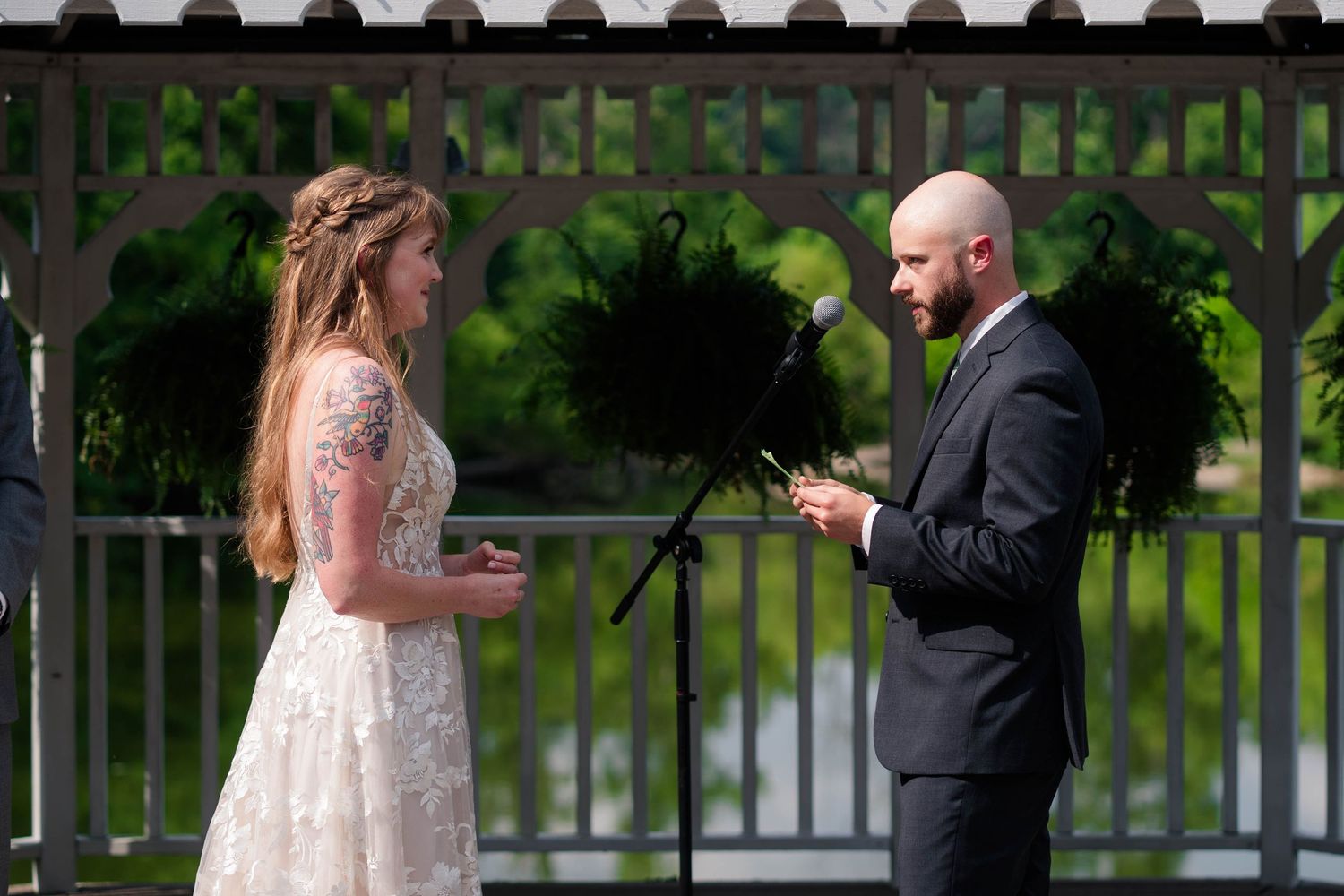 Bride and groom exchange vows at outdoor wedding ceremony near white gazebo on sunny day.