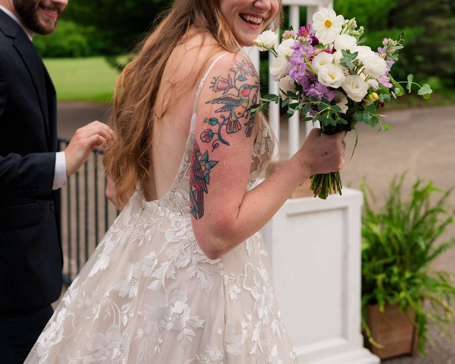 Smiling bride with floral arm tattoo holds colorful bouquet in backless lace wedding gown outdoors.