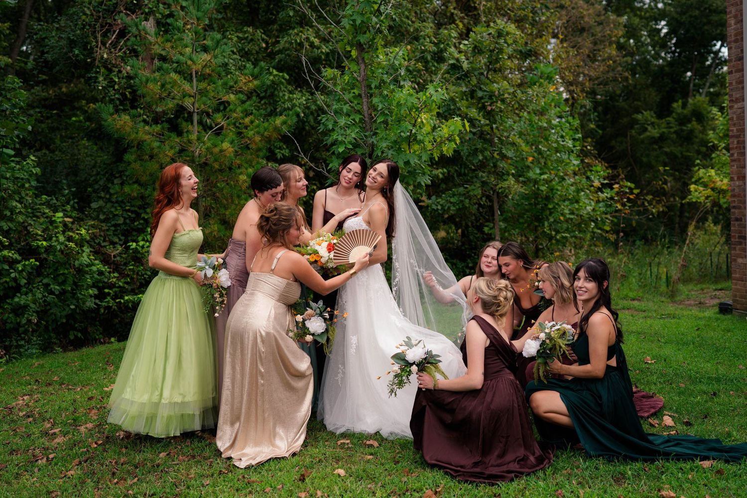 Bride surrounded by bridesmaids in mismatched green, gold, and burgundy gowns on lush garden lawn.