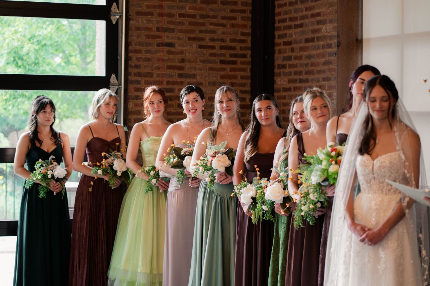 Bridesmaids in mismatched earth-tone gowns hold floral bouquets alongside bride in white wedding dress.