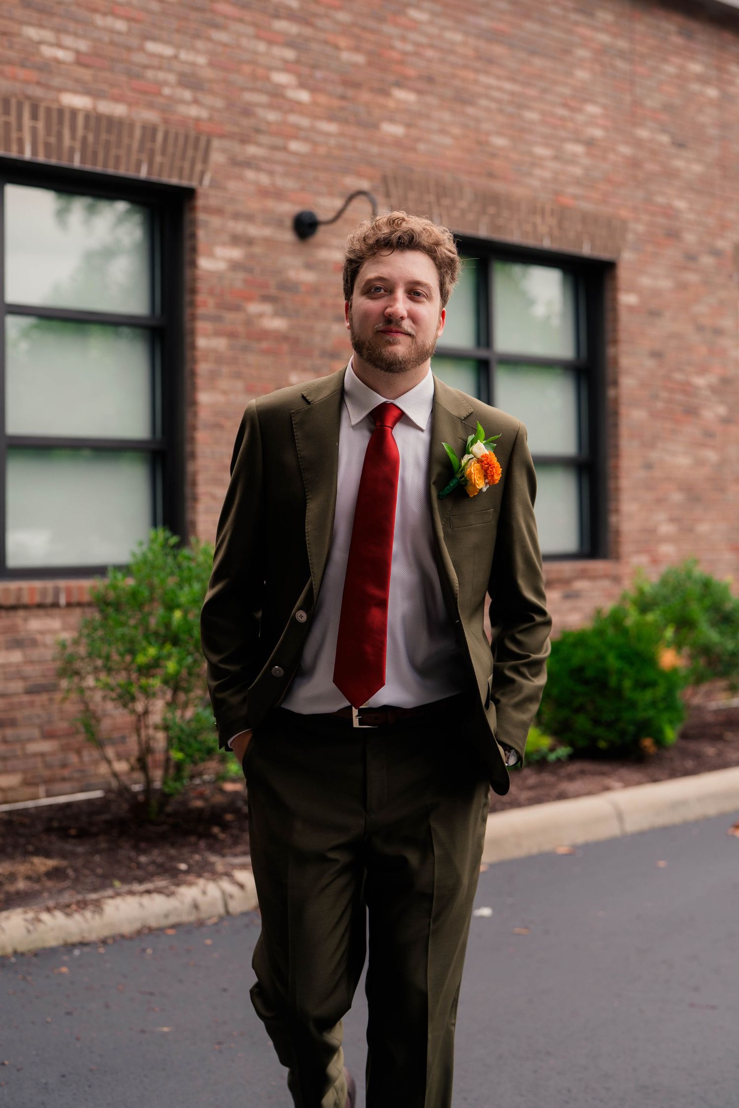 Groom in olive green suit with red tie and orange boutonniere walks outside brick wedding venue.