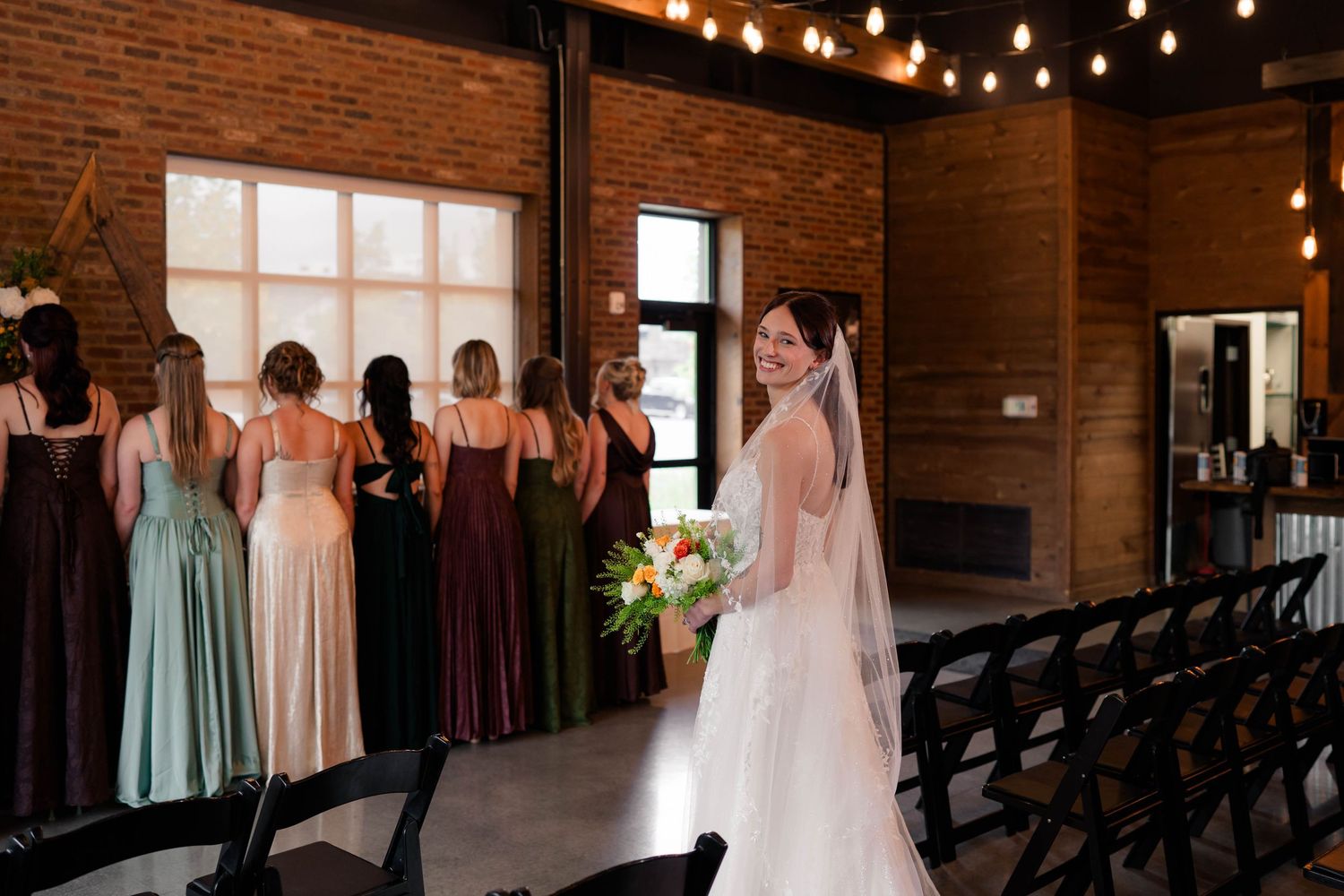 Bride in white gown with veil smiles back at bridesmaids in colorful mismatched dresses in a rustic brick venue.