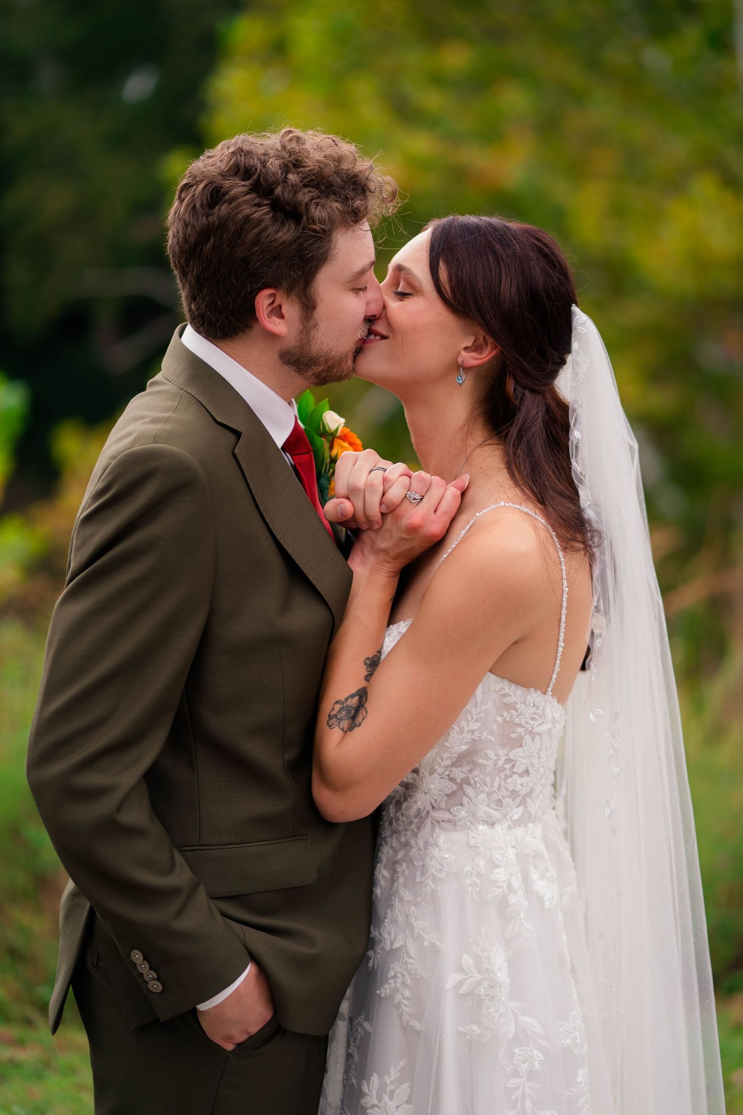 Bride and groom kiss while holding hands outdoors with lush green foliage in the background.