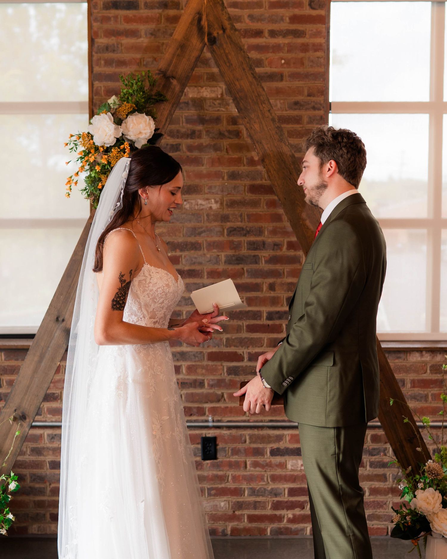 Bride reads vows to groom at rustic brick wall altar with wooden arch and floral arrangements.
