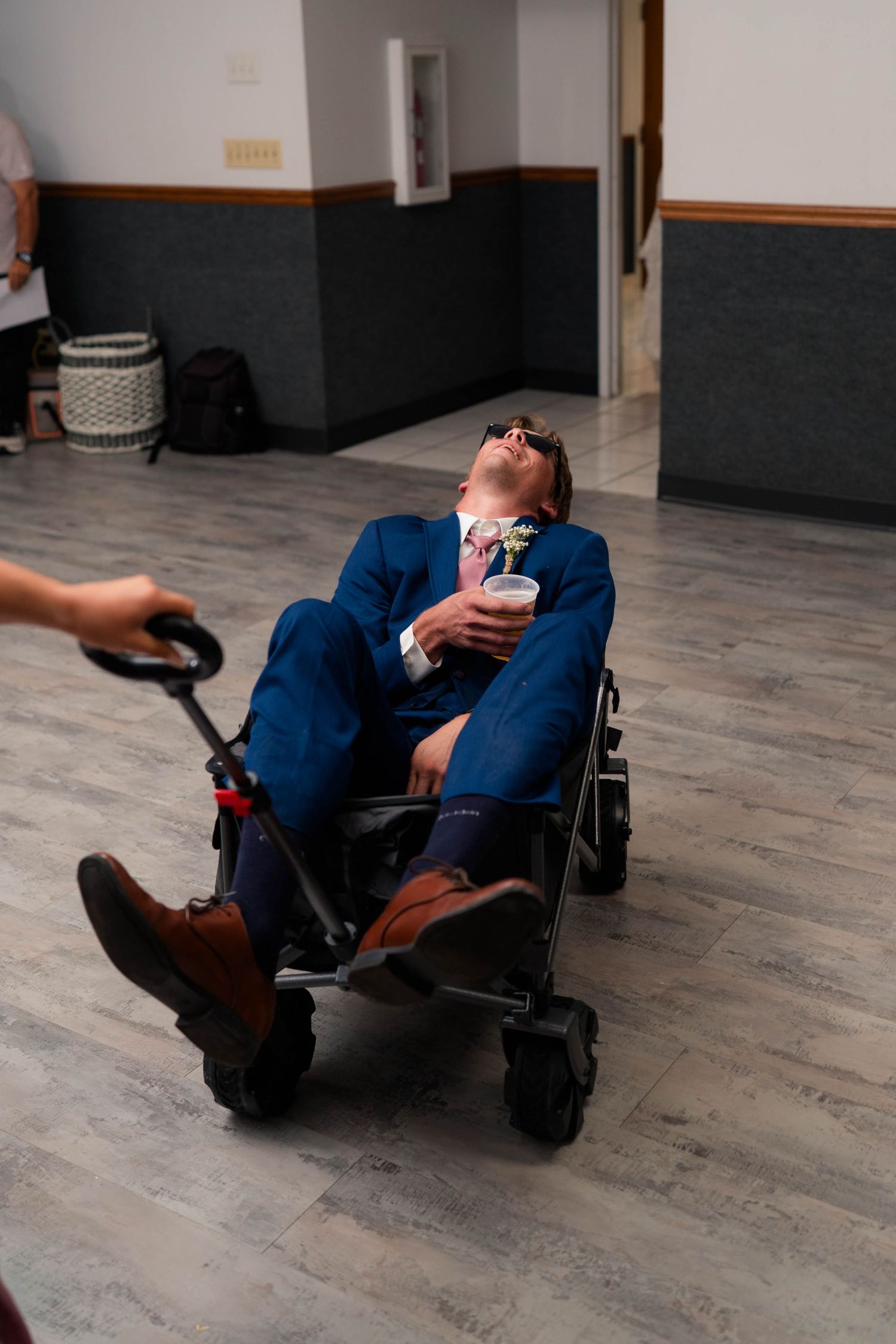 Groom in navy suit reclines in wagon holding drink with head back while being pulled across venue floor.
