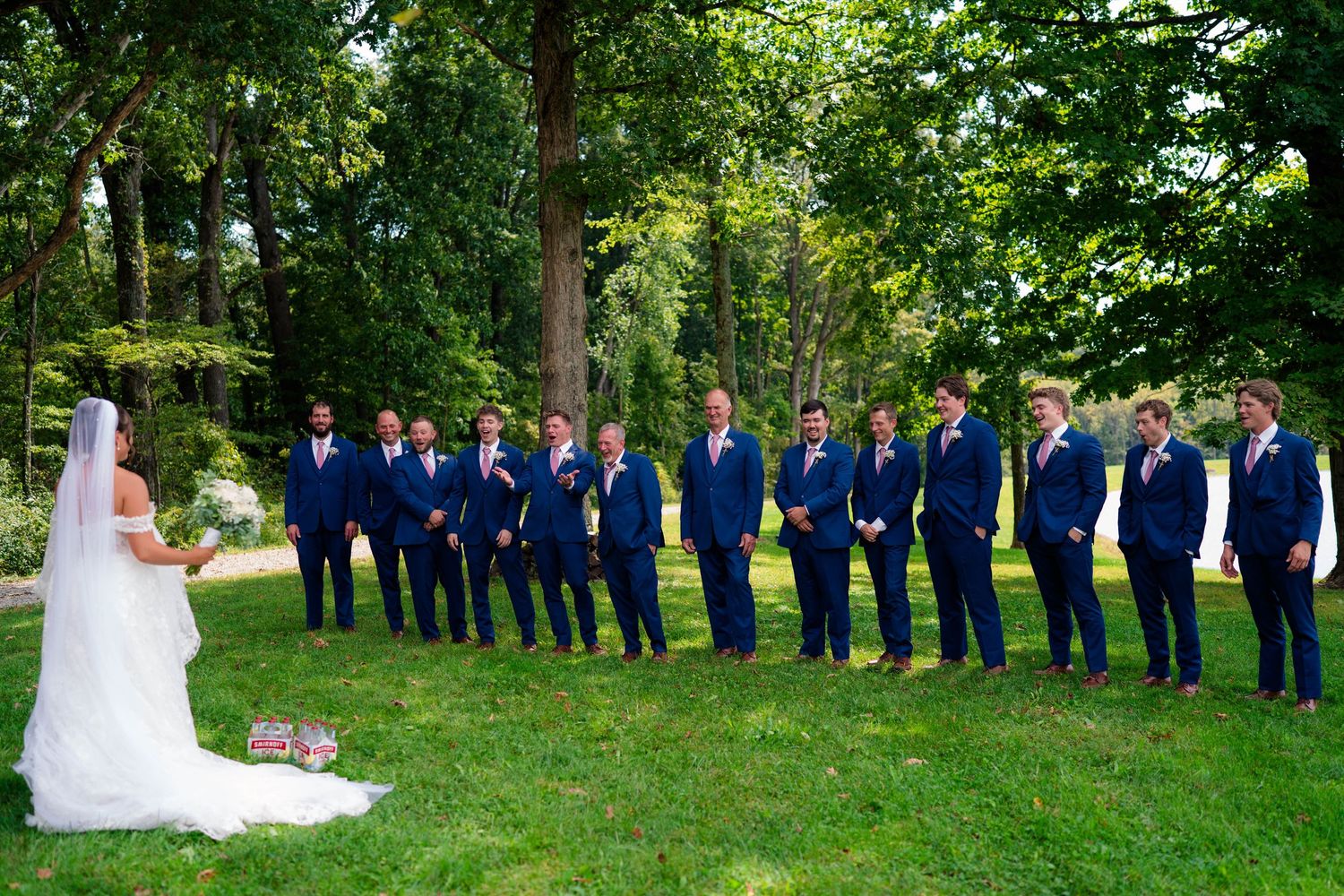 Bride faces a line of groomsmen in navy suits on a lush green lawn during wedding portraits.