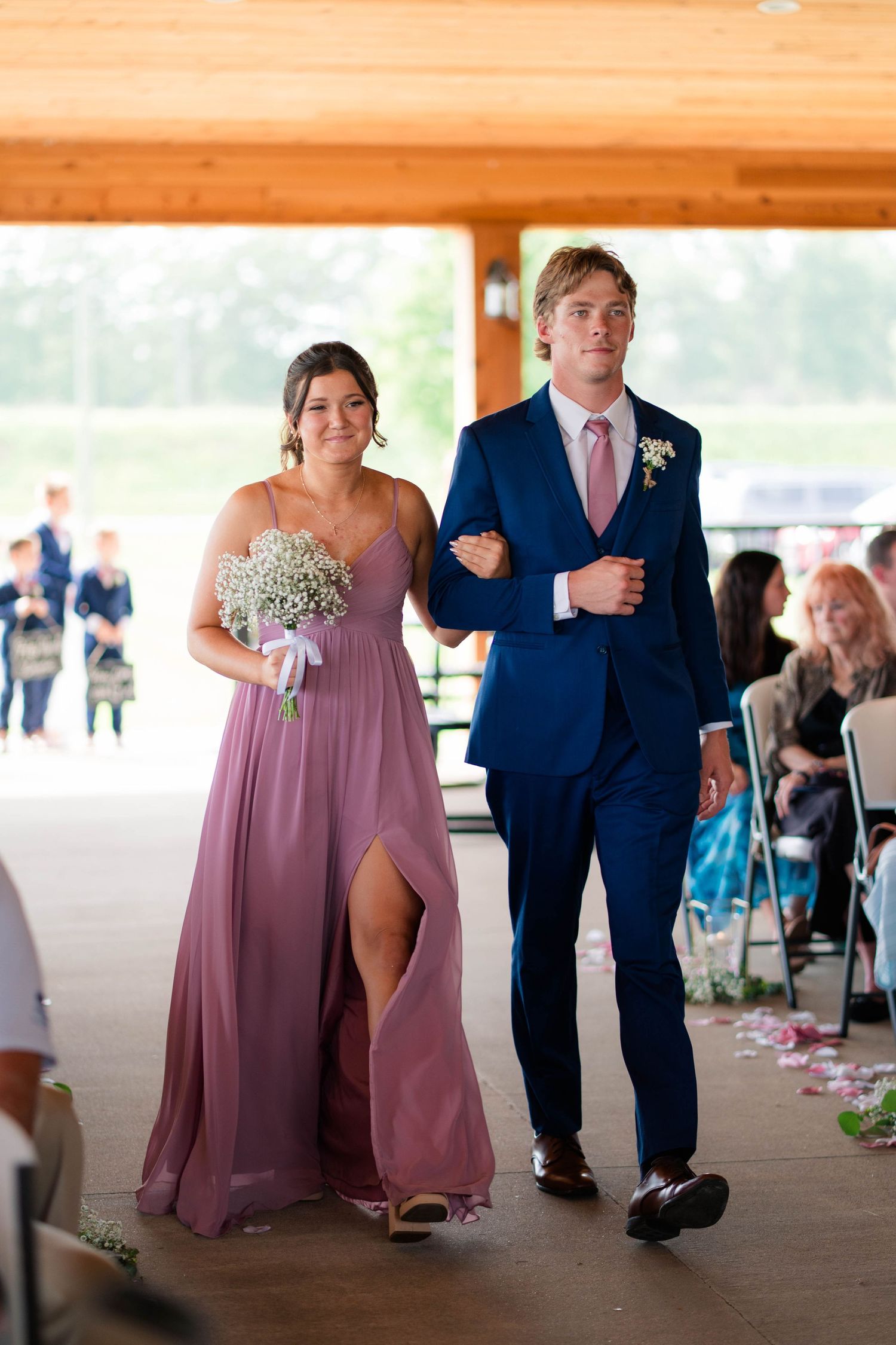 Groomsman in navy suit and pink tie escorts bridesmaid in mauve dress down aisle.