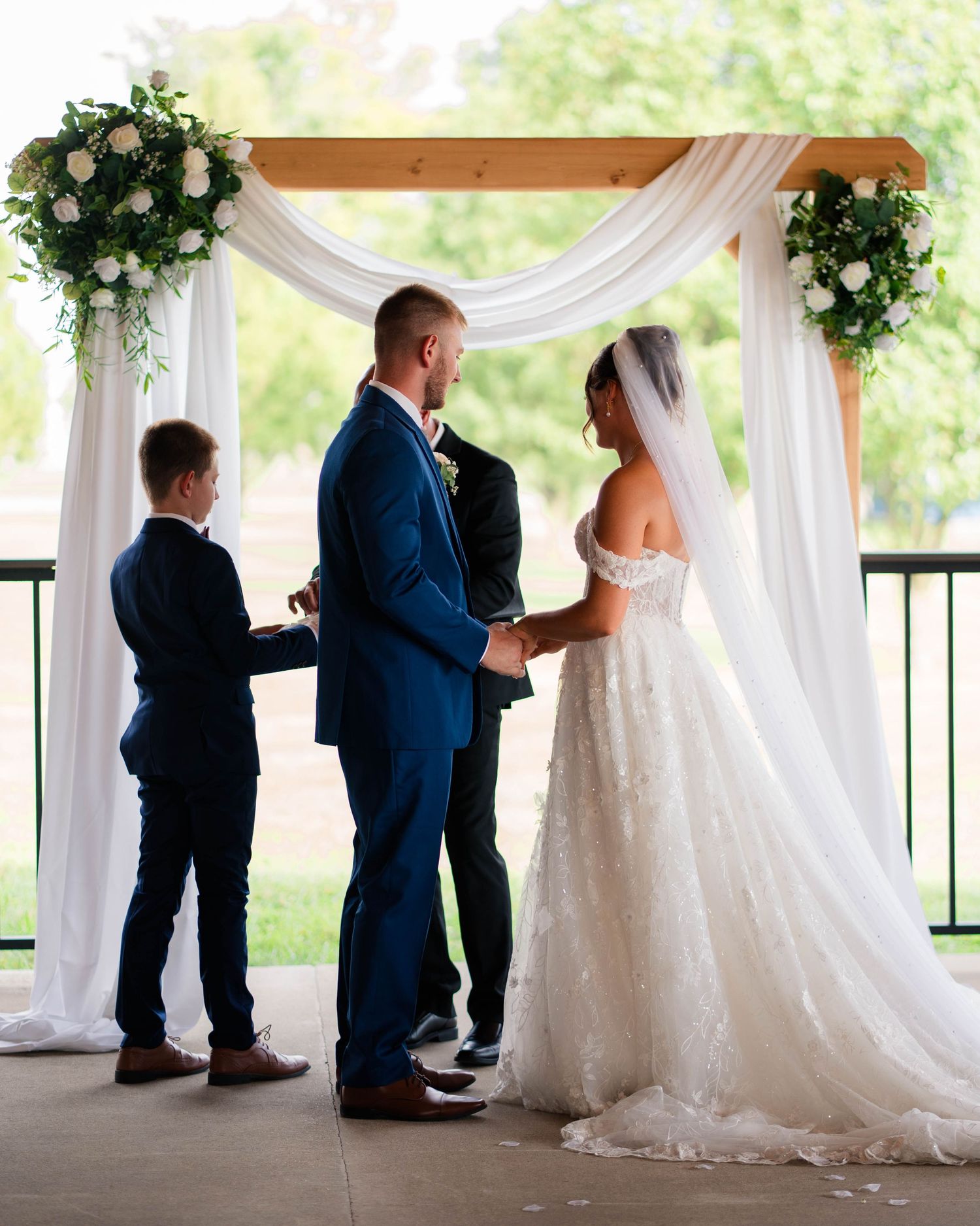 Bride and groom exchange vows under a wooden arch with white draping and floral arrangements.