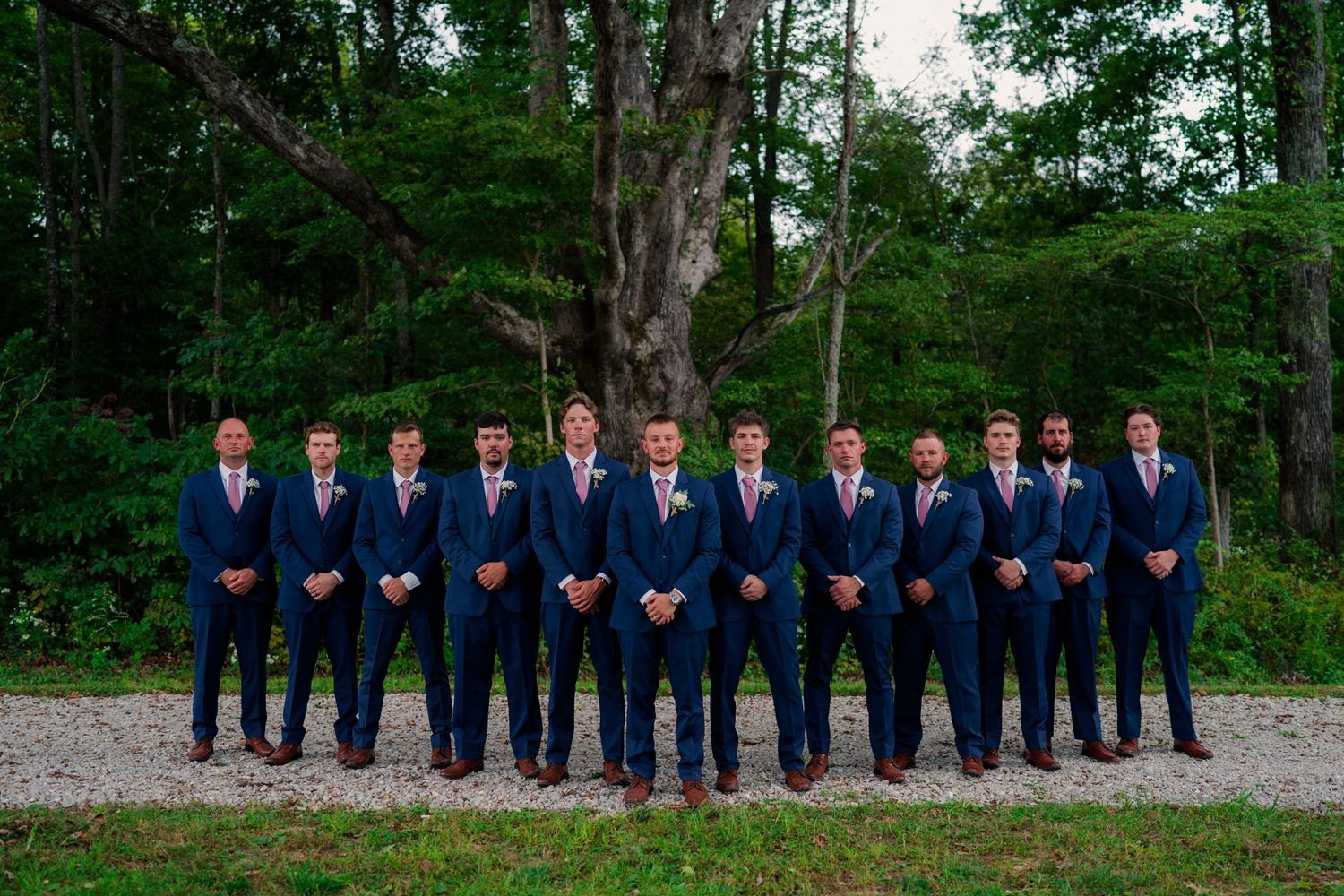 Groomsmen in navy blue suits with pink ties lined up outdoors before a wedding ceremony.