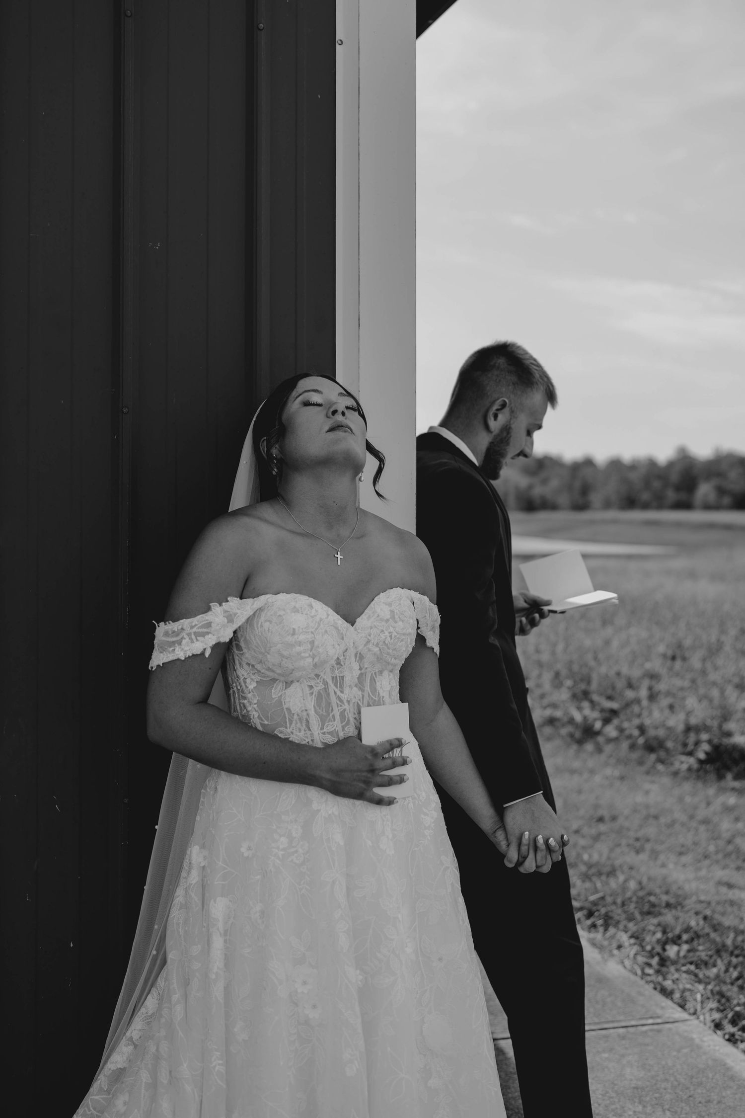 Black and white photo of bride and groom holding hands while privately reading vows outside barn.