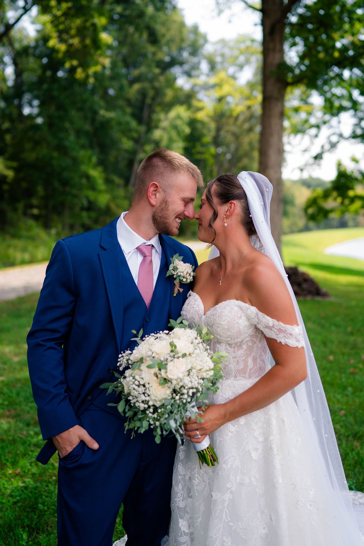 Bride and groom share a romantic moment outdoors, bride holding white bouquet in lace gown with veil.