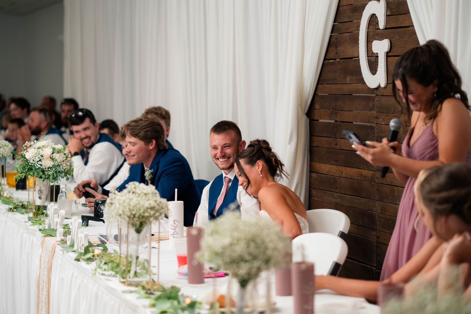 Bride and groom laugh together at decorated head table during wedding reception with guests in background.