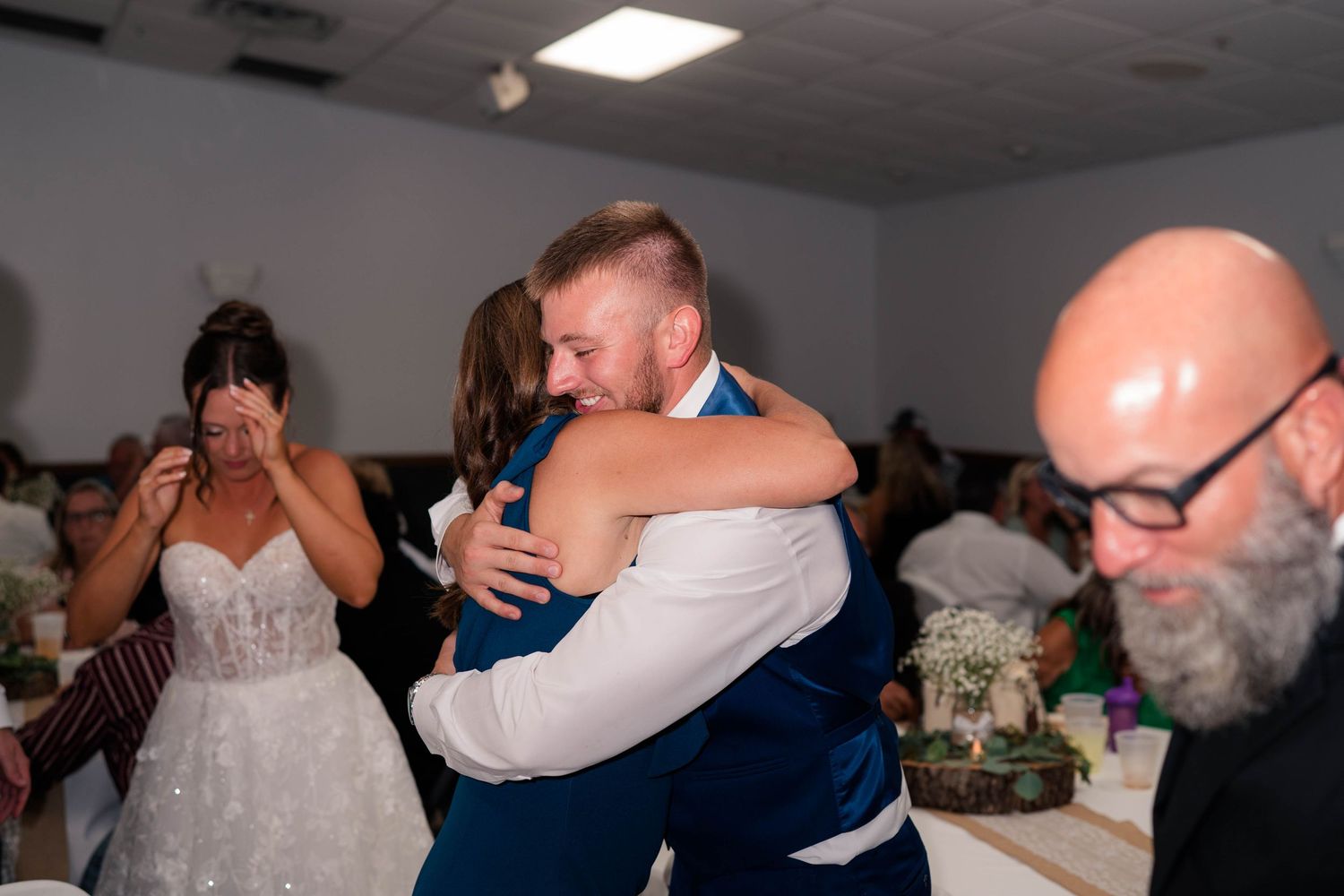 Groom hugging a guest at wedding reception while emotional bride stands nearby in white dress.