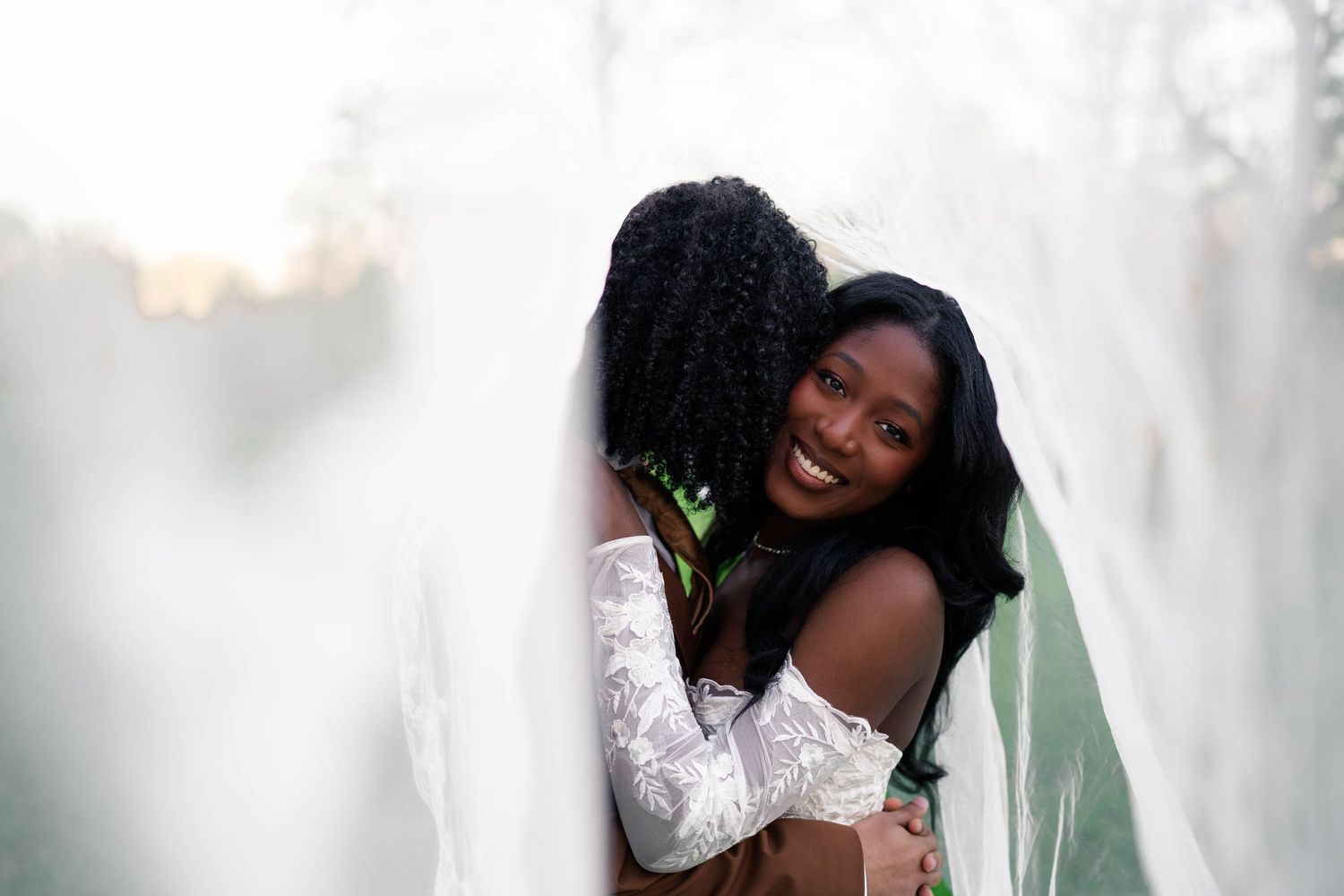 Two Black women sharing a joyful hug outdoors, one wearing a white lace wedding dress and veil.