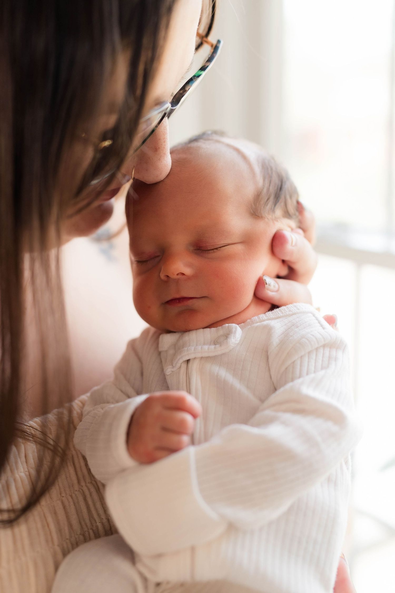 Mother tenderly holds sleeping newborn baby dressed in white against her chest near a bright window.