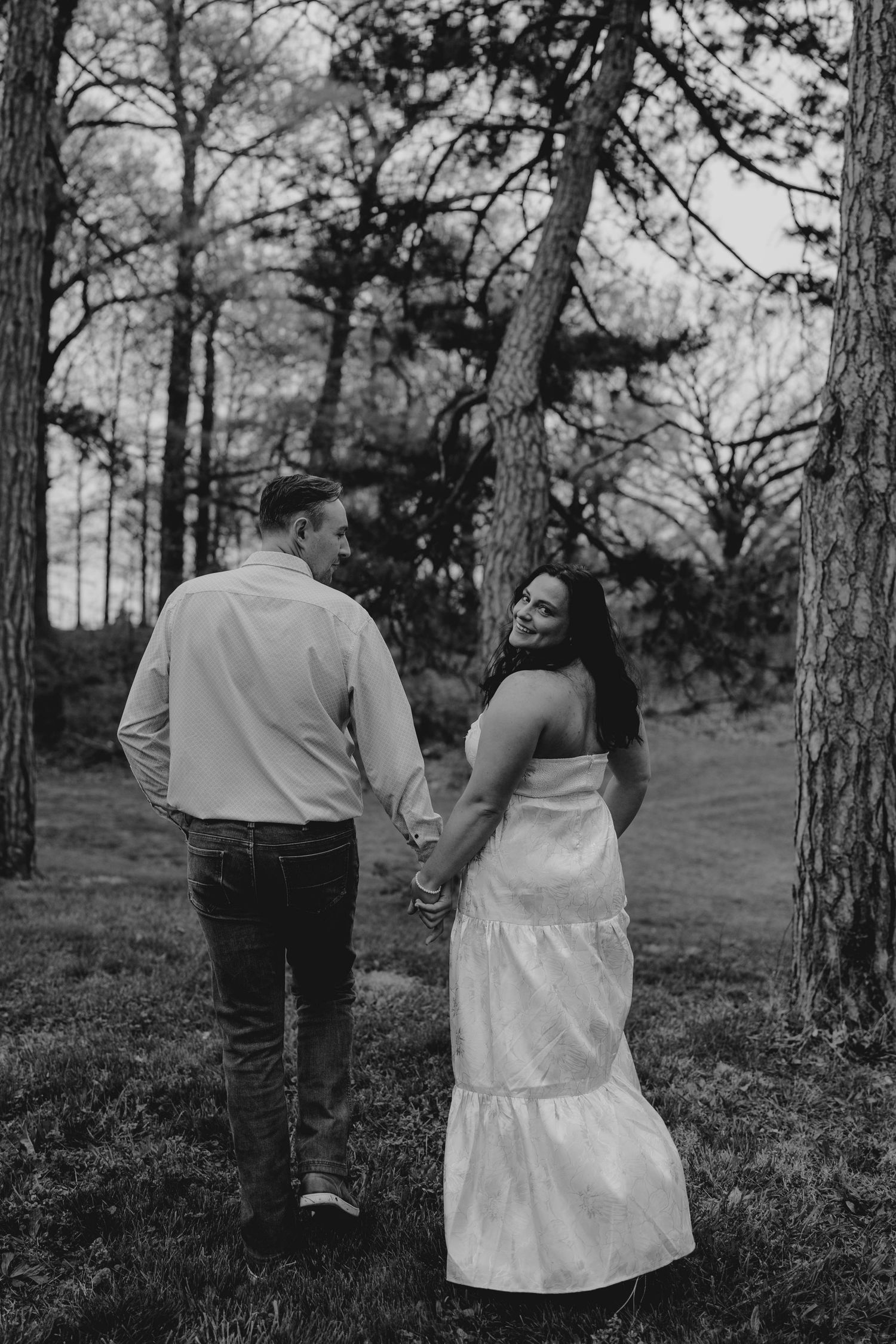 Black and white photo of couple holding hands walking through pine forest, woman looking back.