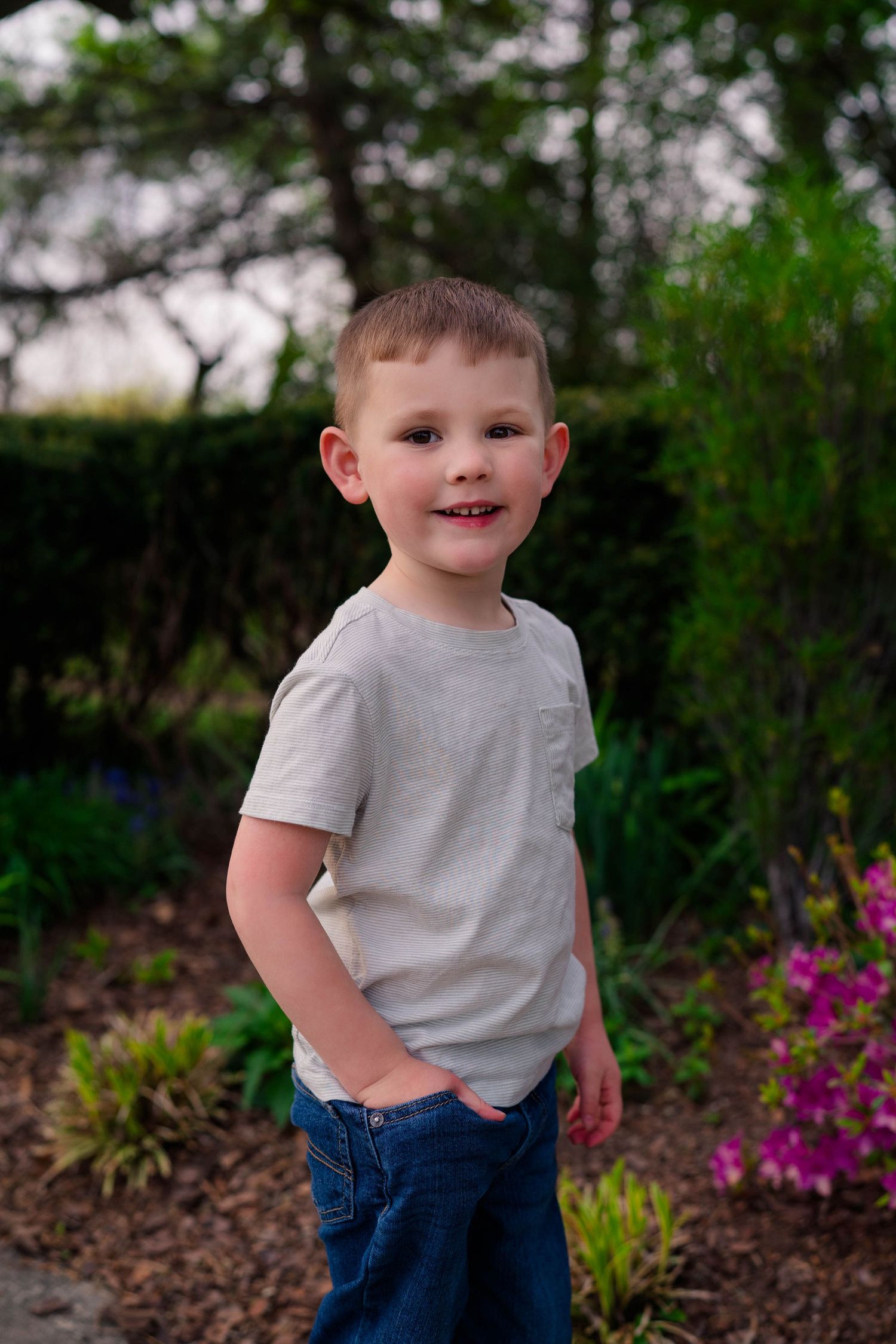 Young boy in gray t-shirt and jeans smiling in a lush garden setting.