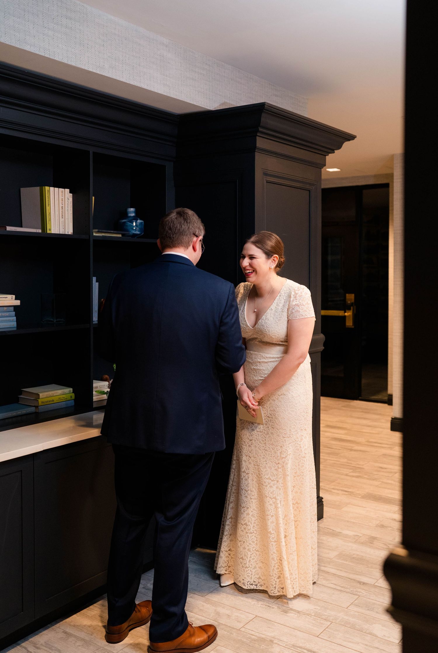 Smiling bride in lace gown holds grooms hands during an emotional first look reveal in hotel.