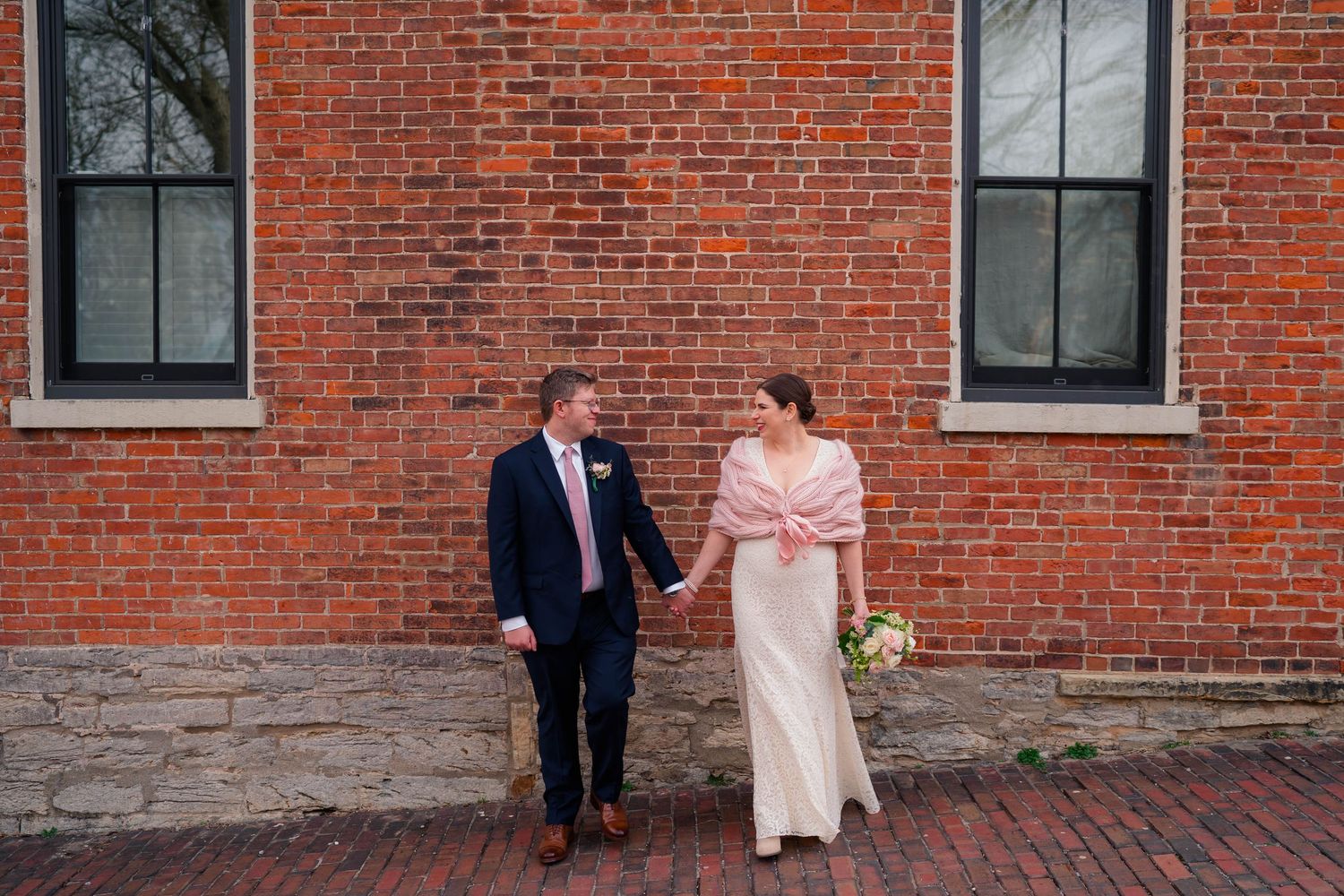 Bride and groom walk hand-in-hand along a brick path beside a historic red brick building.