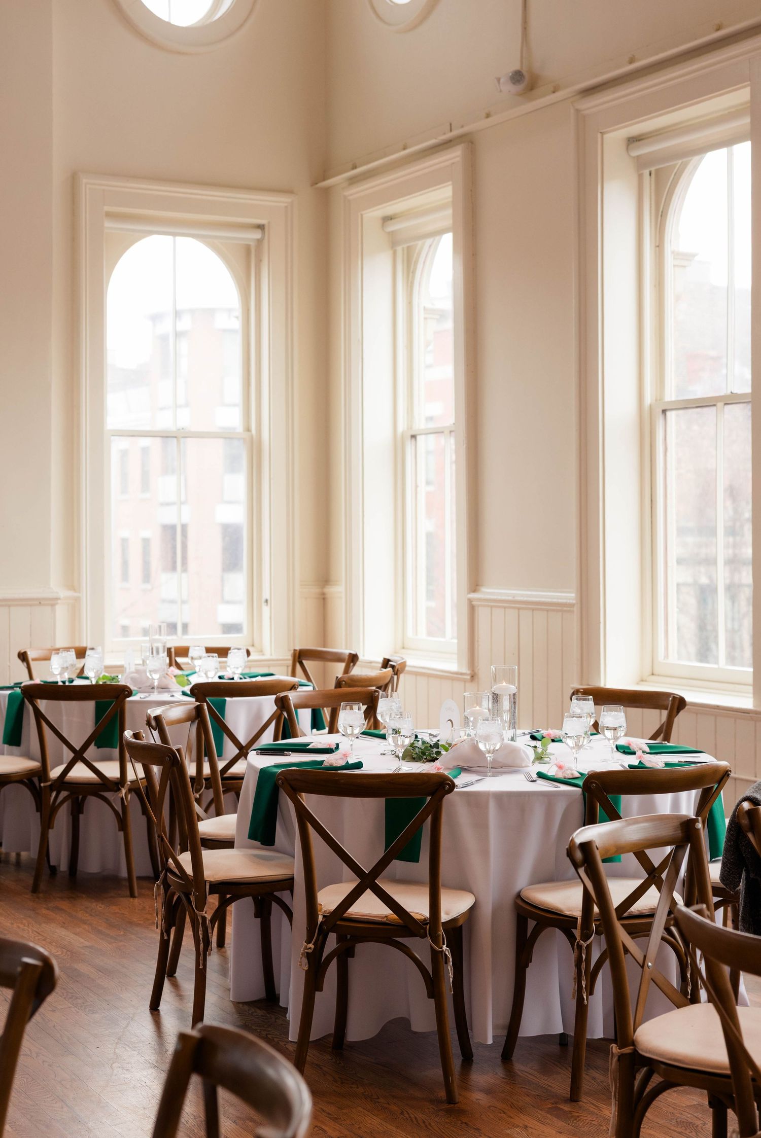 Bright wedding reception room with arched windows, round tables, emerald green napkins, and wooden cross-back chairs.