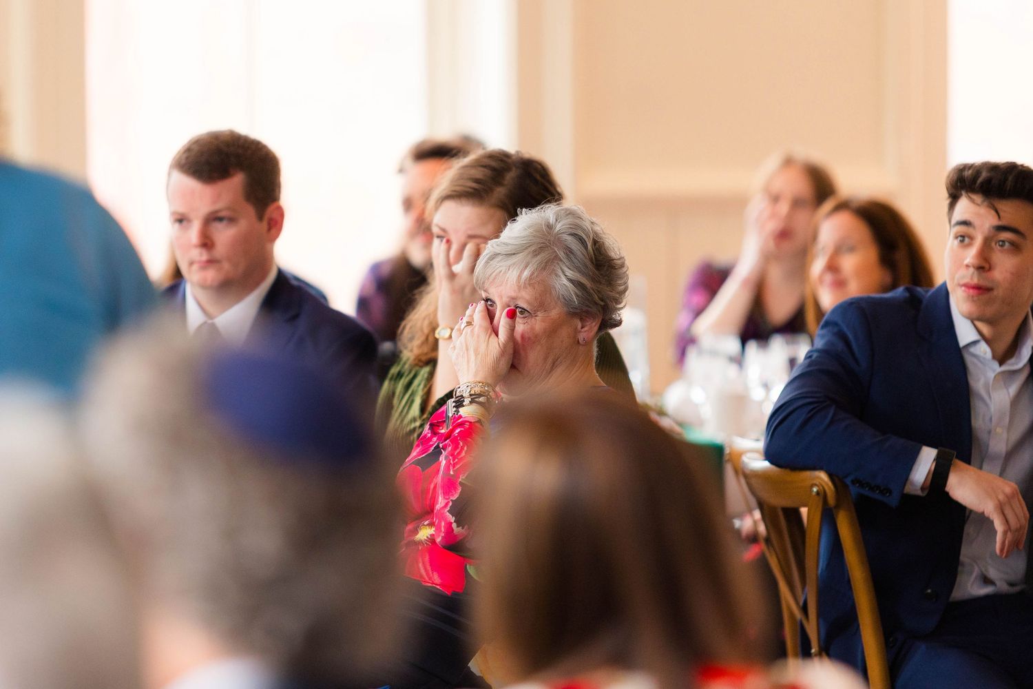 Emotional wedding guest covers face with hands while watching ceremony among seated guests.
