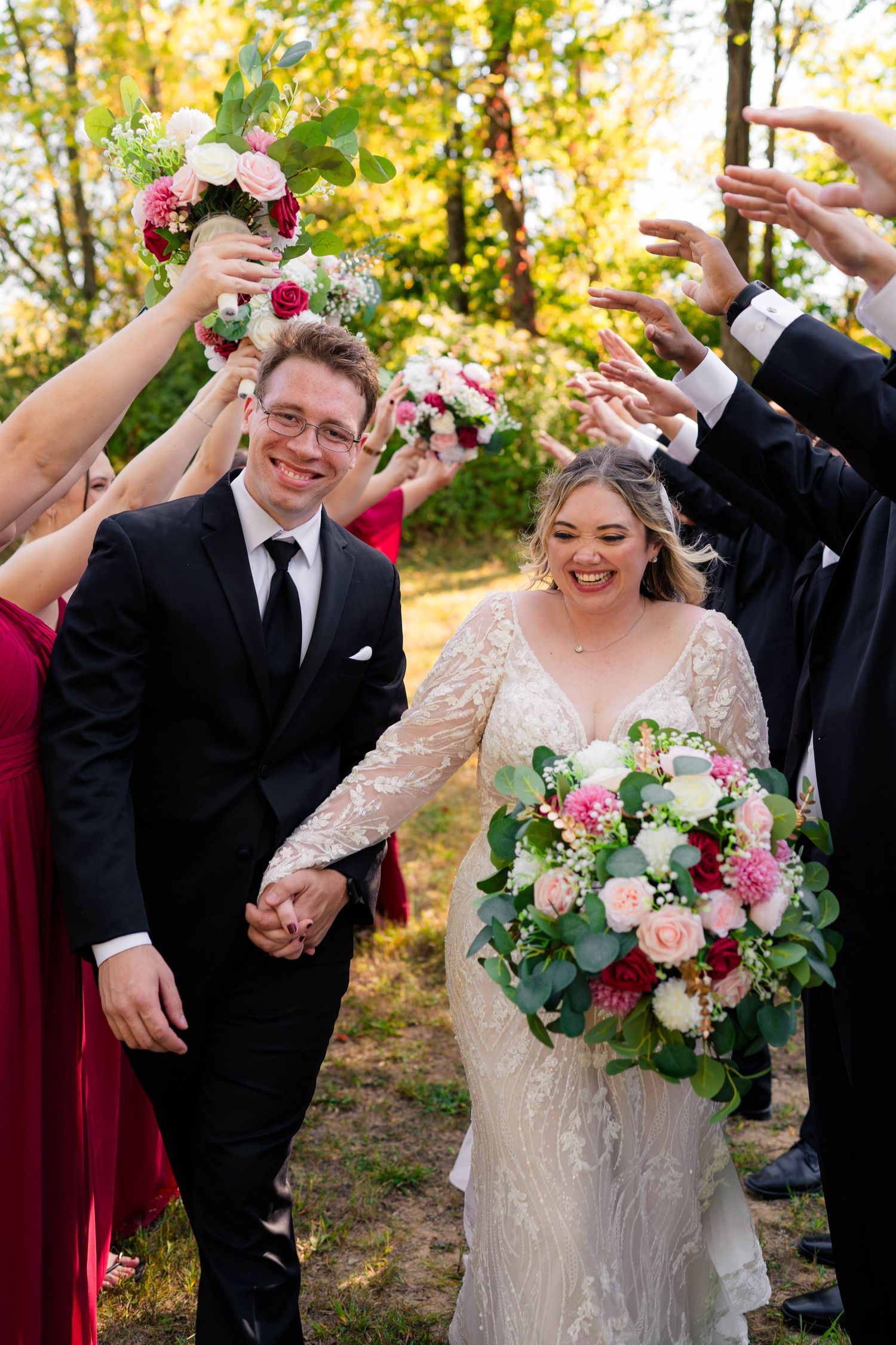 Joyful newlyweds walk hand-in-hand through a tunnel of raised bouquets, bride in lace gown holding pink floral arrangement.