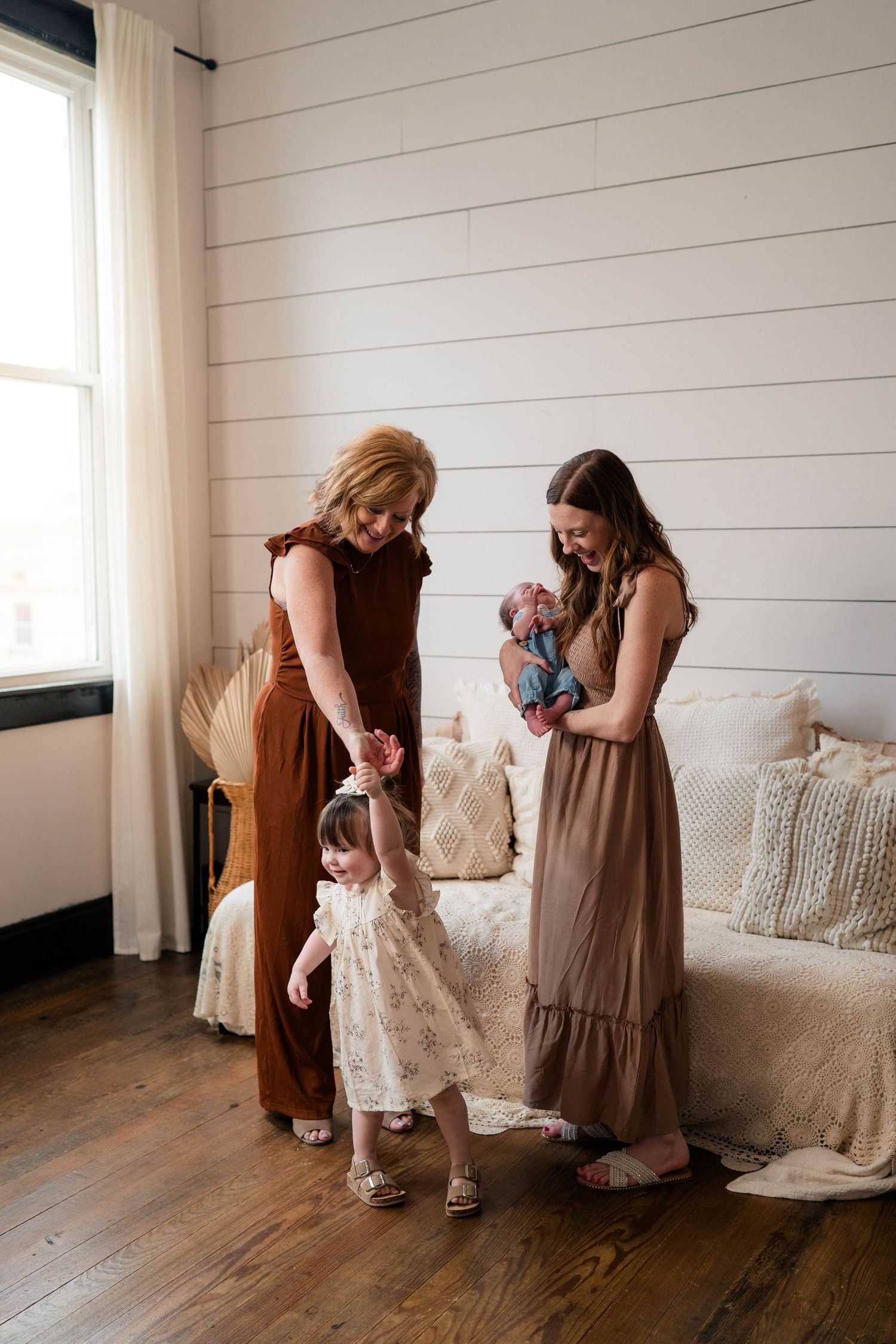 Three generations of women in boho home, grandmother holding toddler's hand while mother cradles newborn.