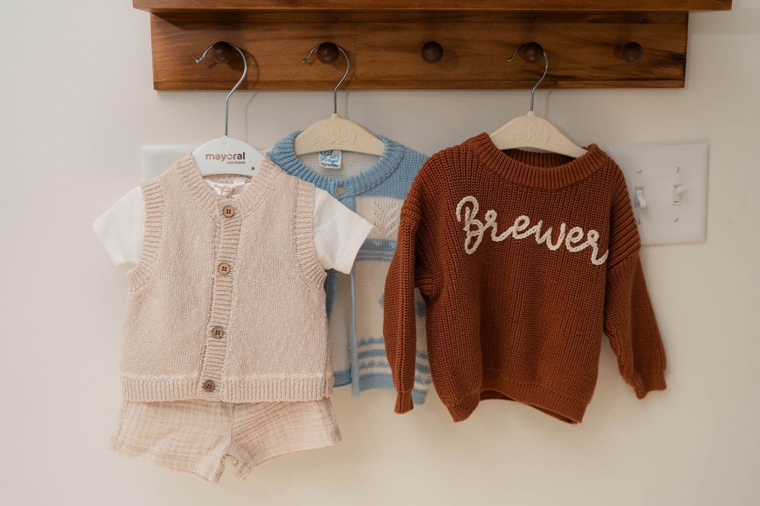 Three baby outfits hang on wooden hooks, including a personalized brown knit Brewer sweater.
