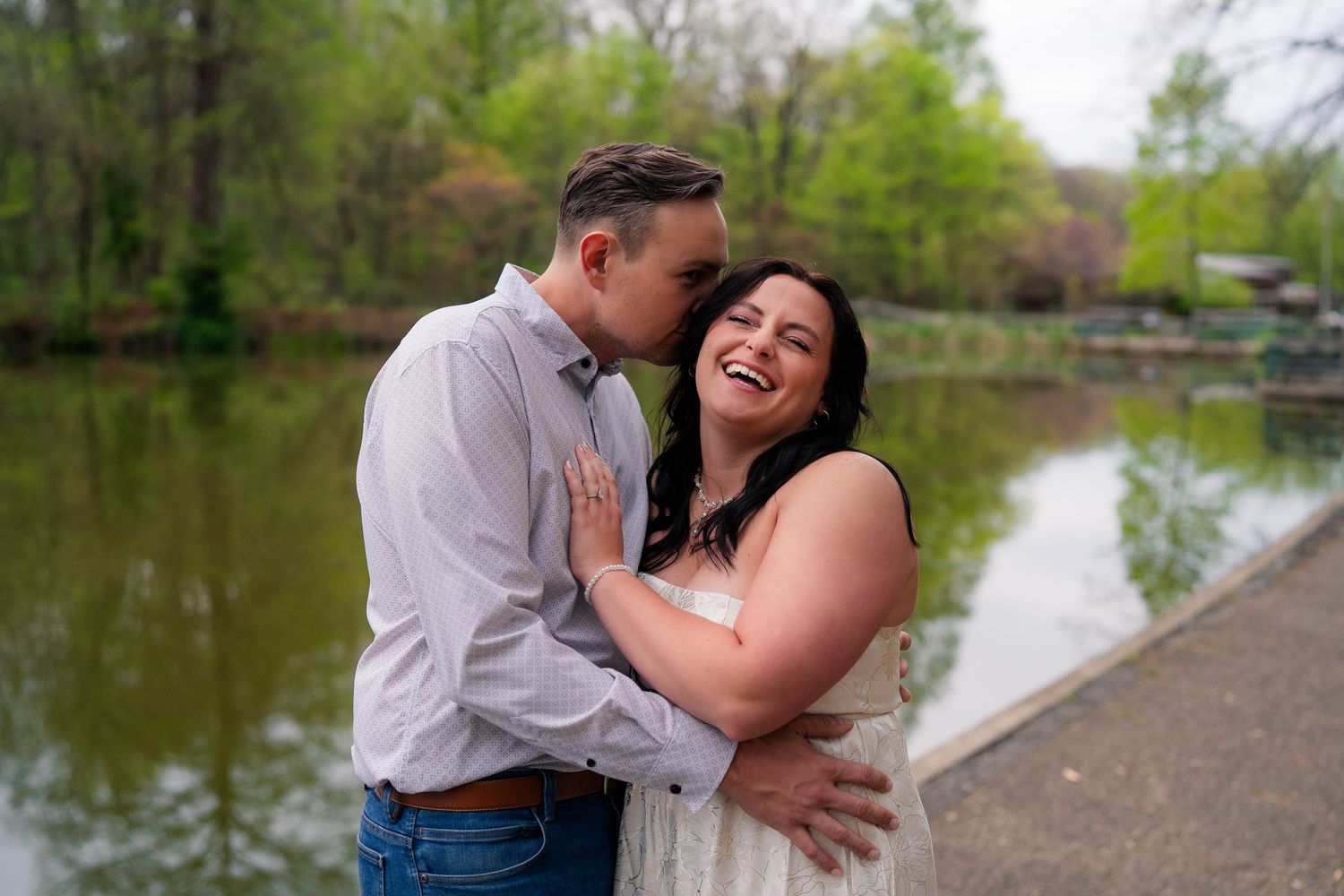 Laughing bride in white dress embraced by groom in lavender shirt near a serene pond during spring engagement photos.