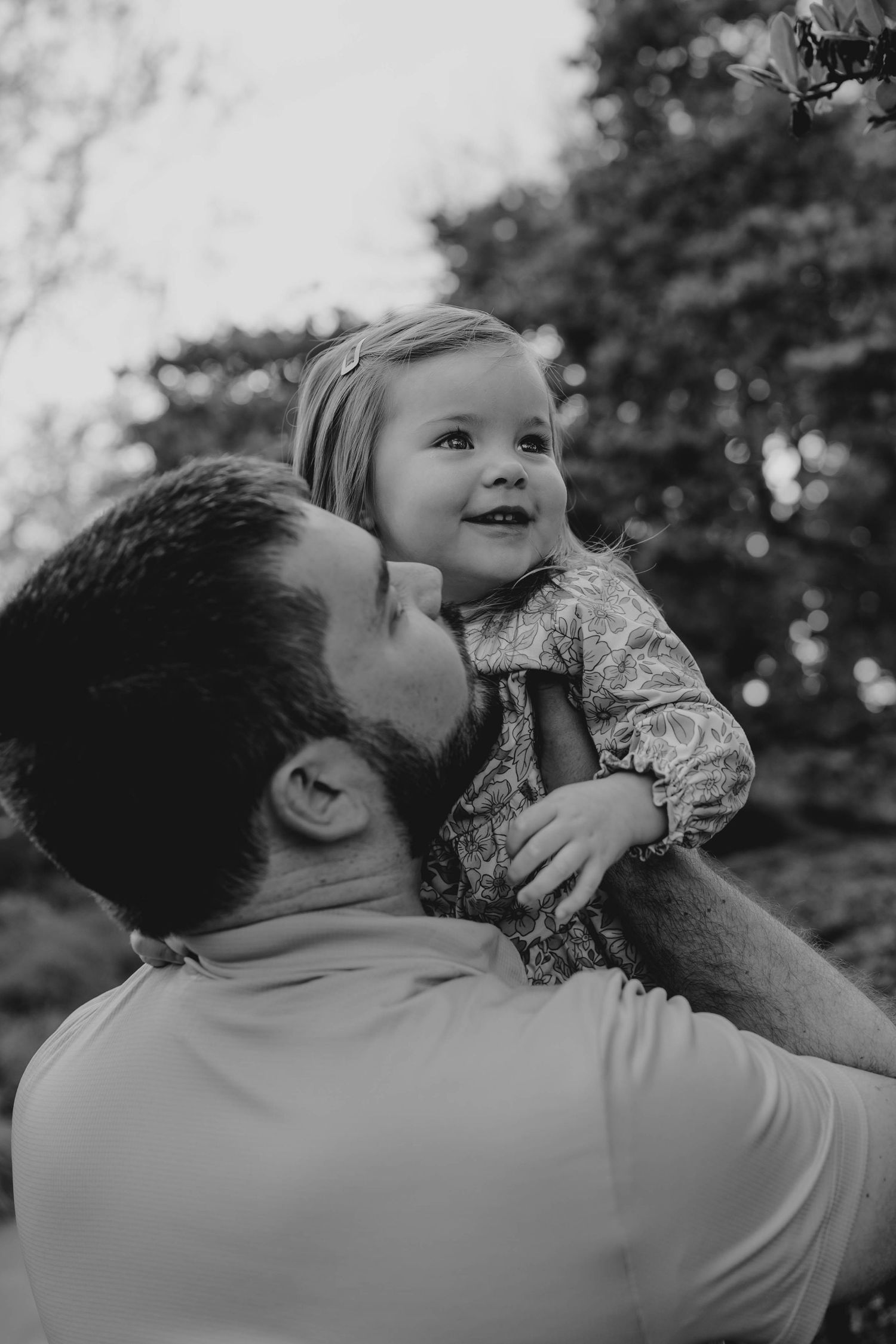 Black and white photo of a father lifting his smiling toddler daughter outdoors with trees in the background.
