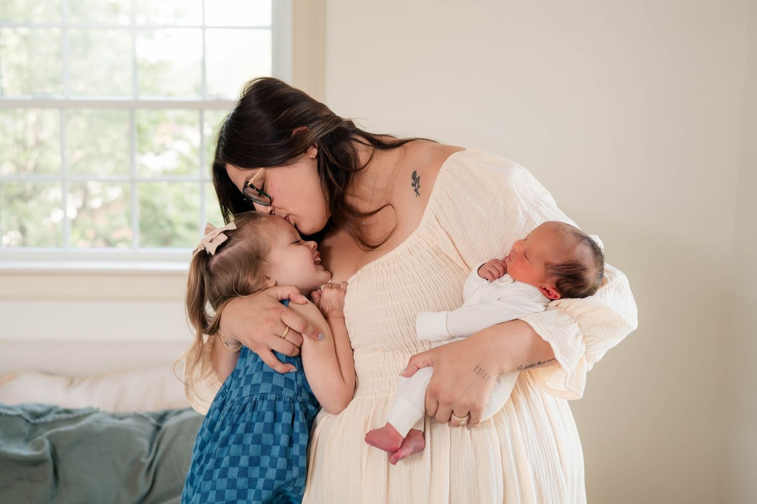 A mother in a white dress holds a newborn while kissing her young daughter in a blue dress near a bright window.