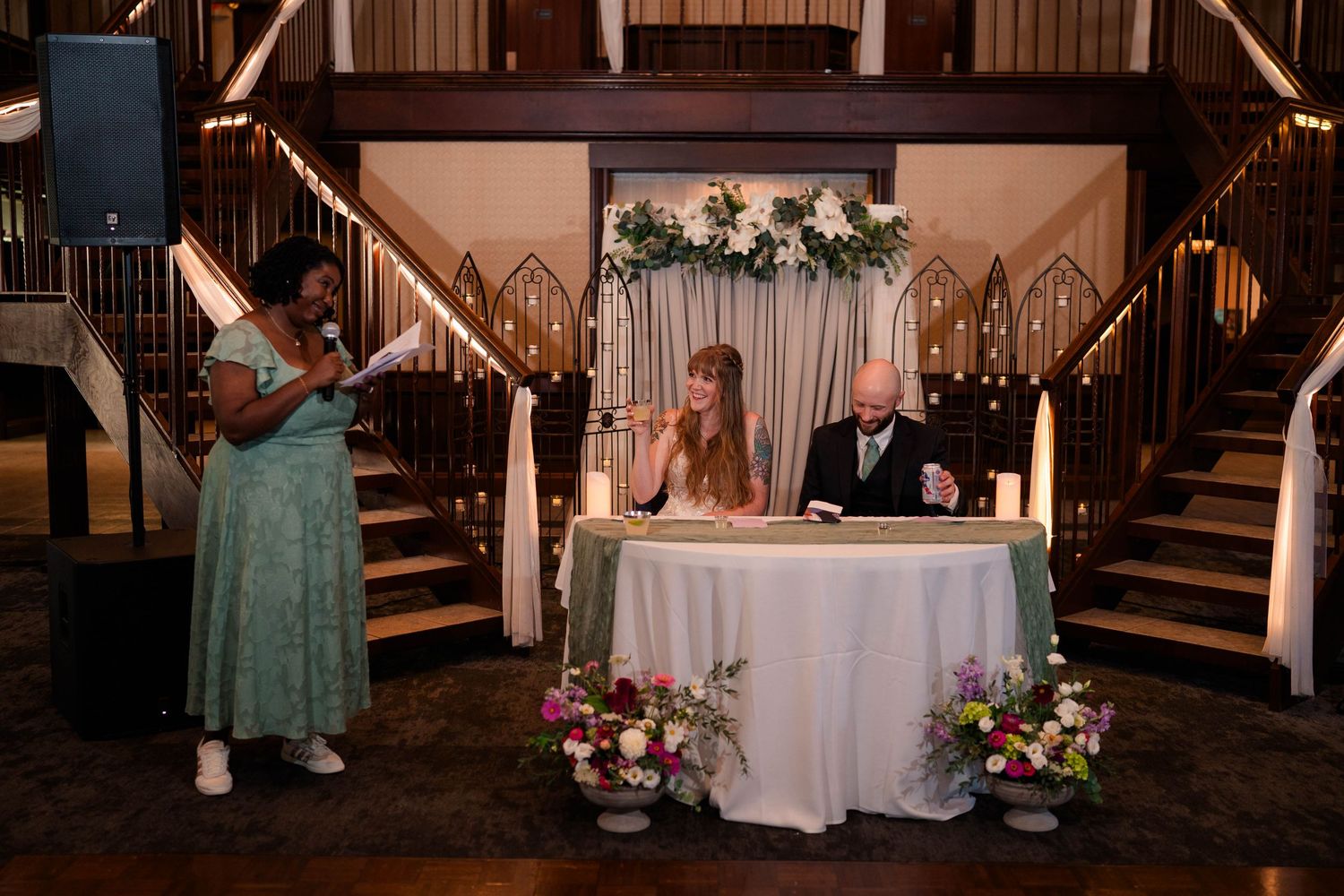 Wedding officiant speaks at microphone while bride and groom sit at sweetheart table.