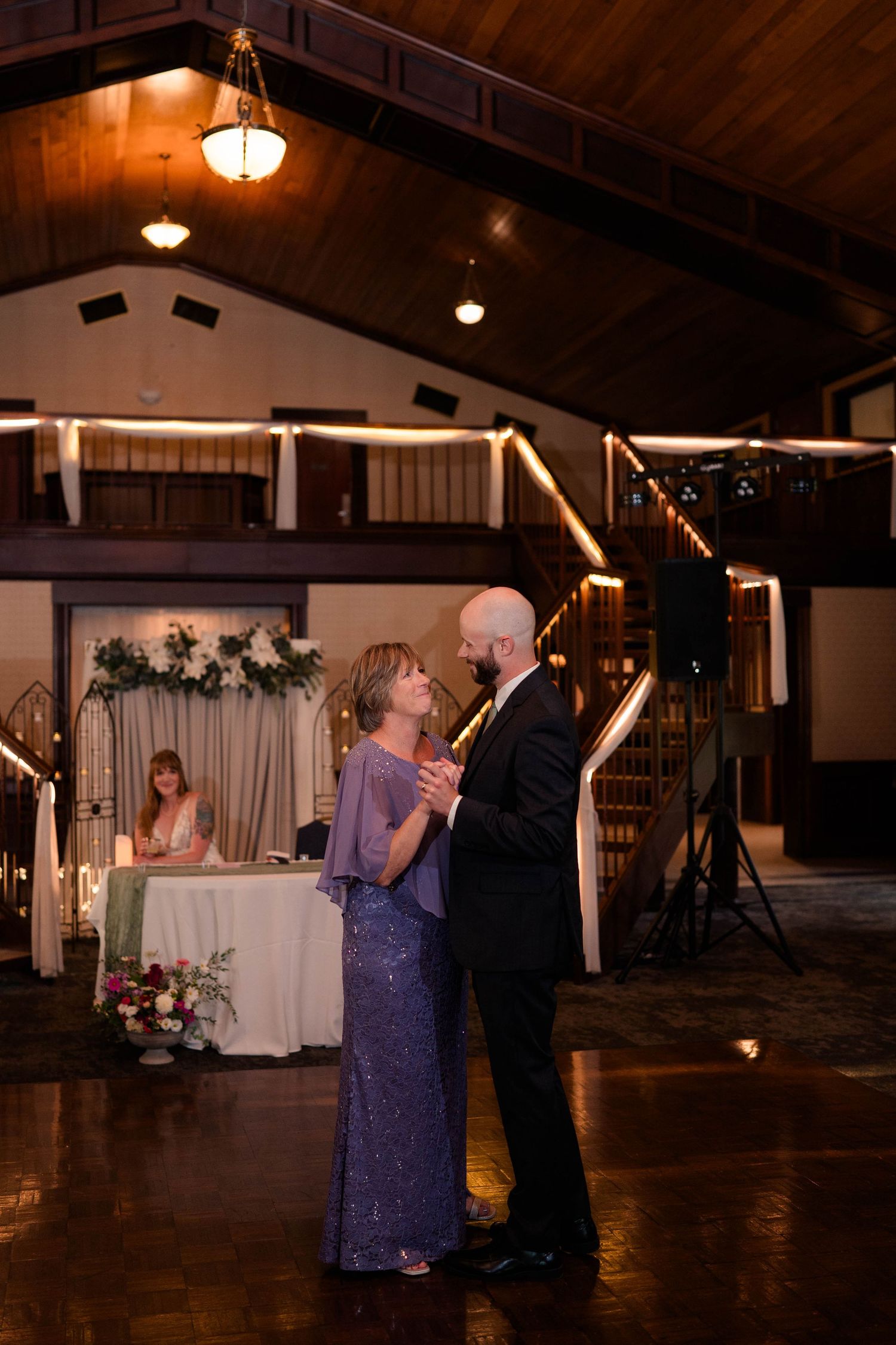 Groom dances with mother in purple sequin dress at wedding reception while bride sits at head table.