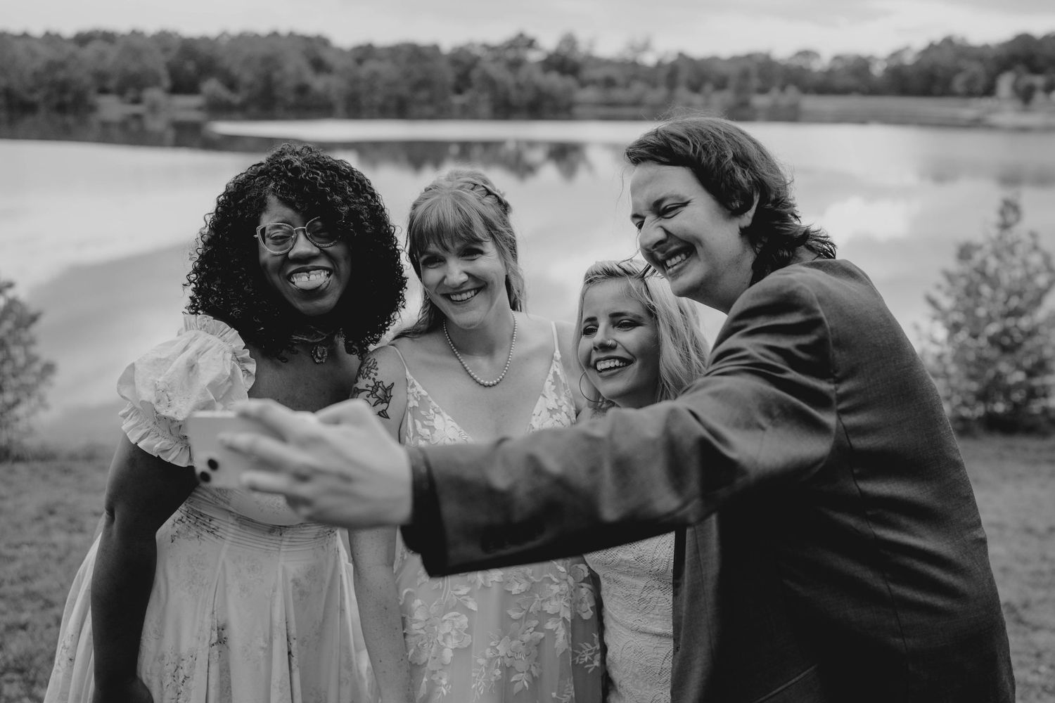 Wedding party takes joyful selfie outdoors near lake in black and white photo.