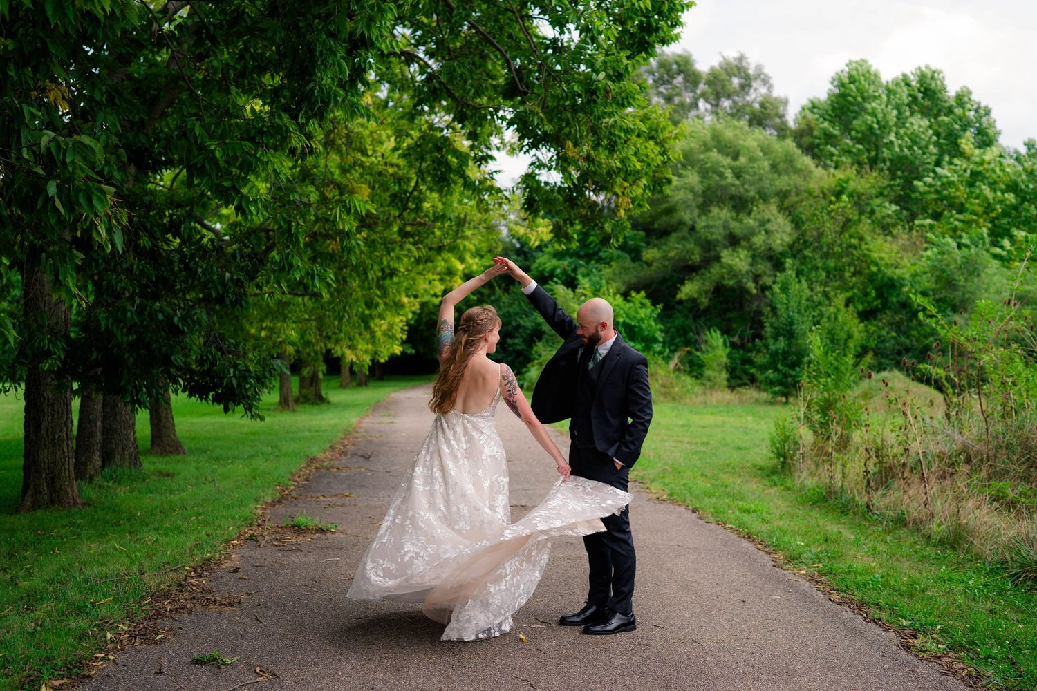 Bride and groom dancing on a tree-lined path in lush green park during outdoor wedding celebration.
