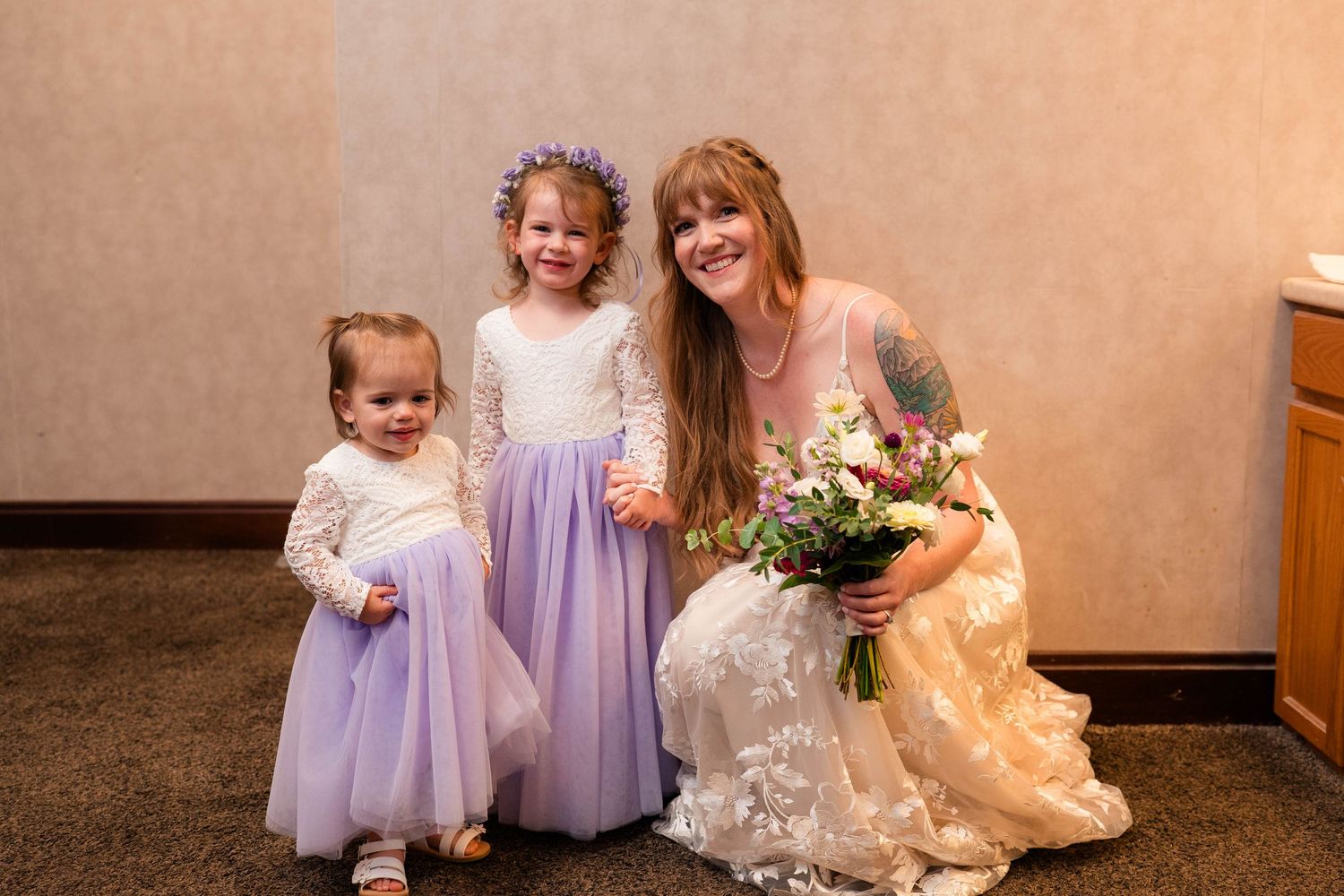 Bride in lace gown kneels beside two flower girls in purple tulle skirts, holding a floral bouquet.