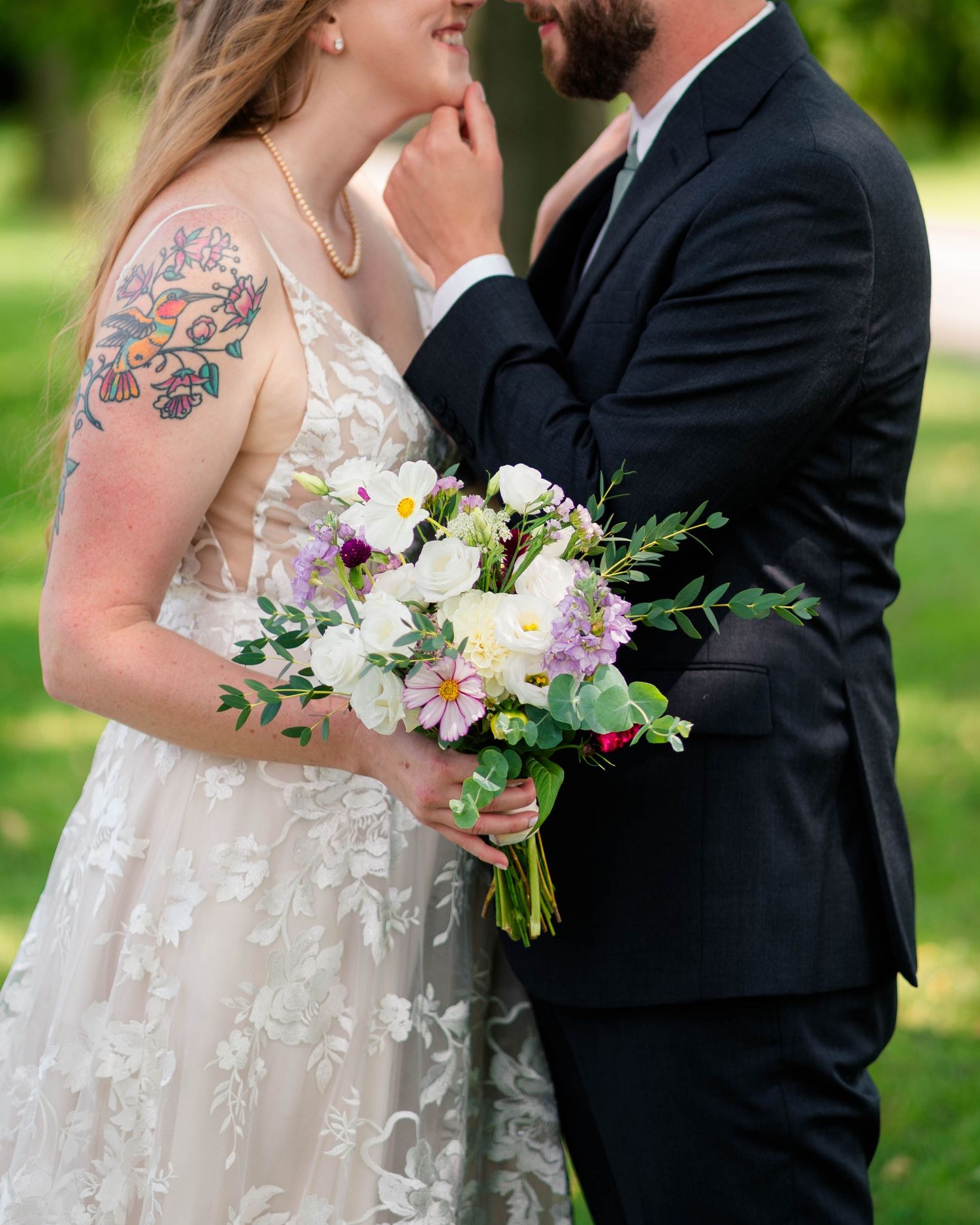 Tattooed bride holds colorful wildflower bouquet while groom gently touches her chin.