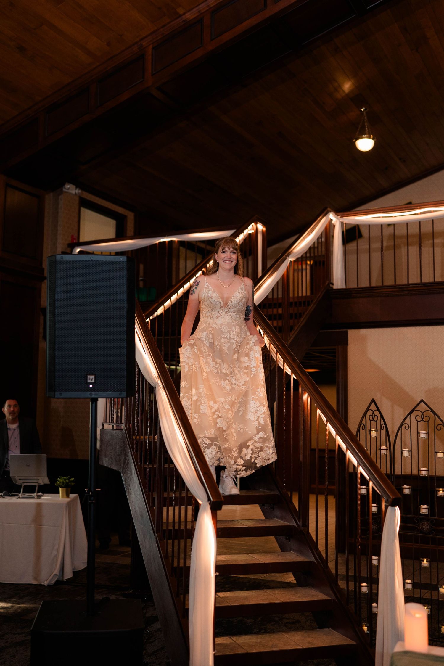 Bride in floral lace gown descends grand staircase at rustic wedding reception venue.