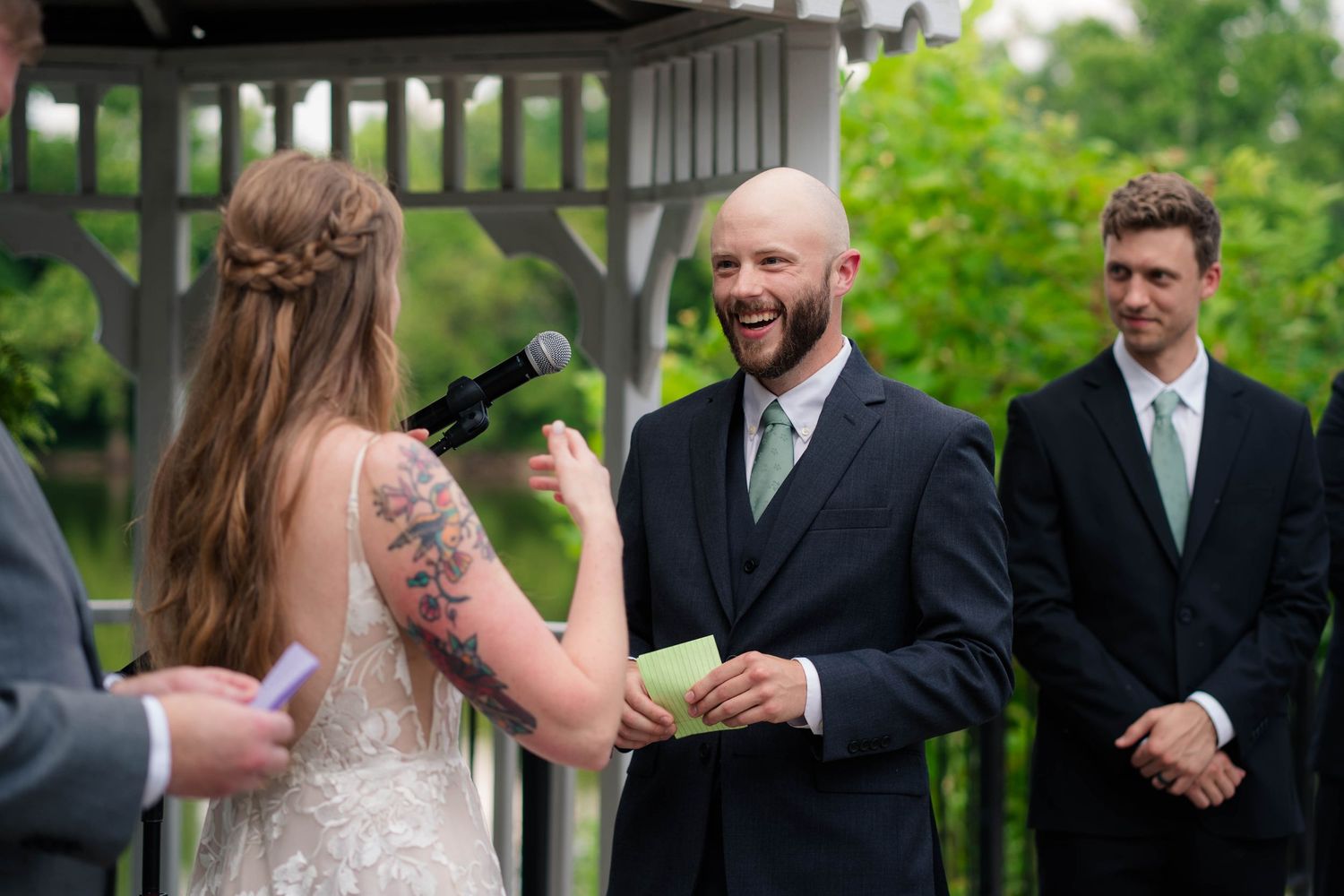 Bald groom laughs joyfully as bride reads wedding vows at outdoor garden ceremony.