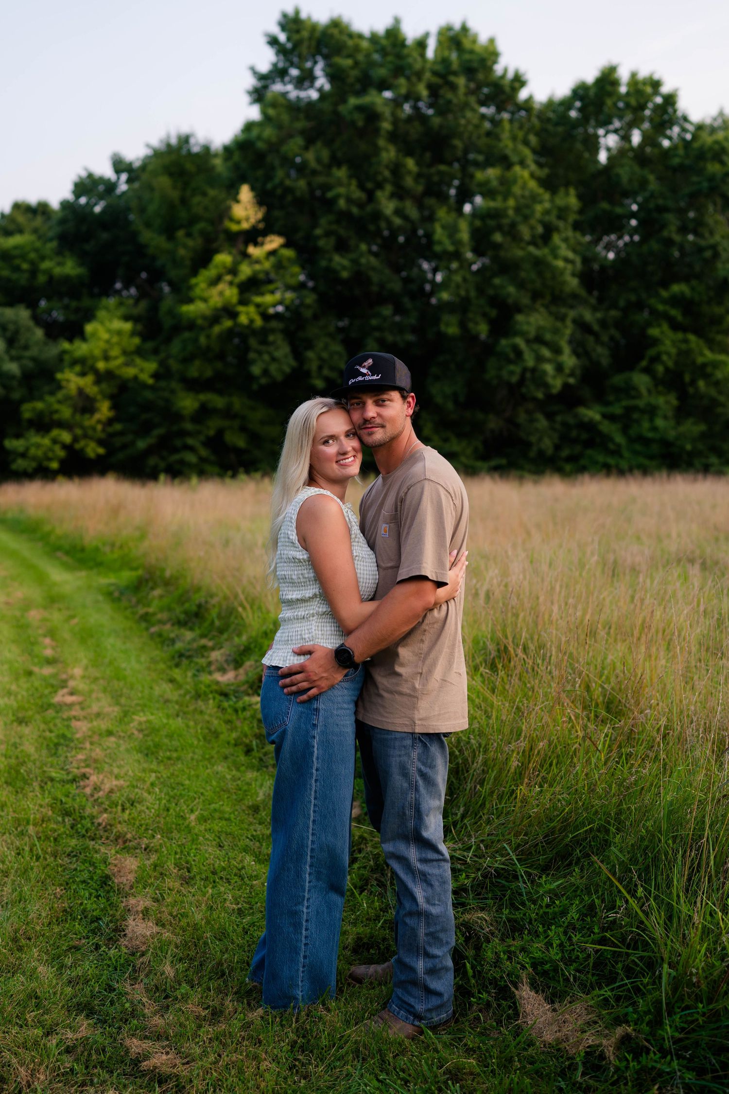 Happy couple embracing and smiling together along a grassy field path at golden hour.