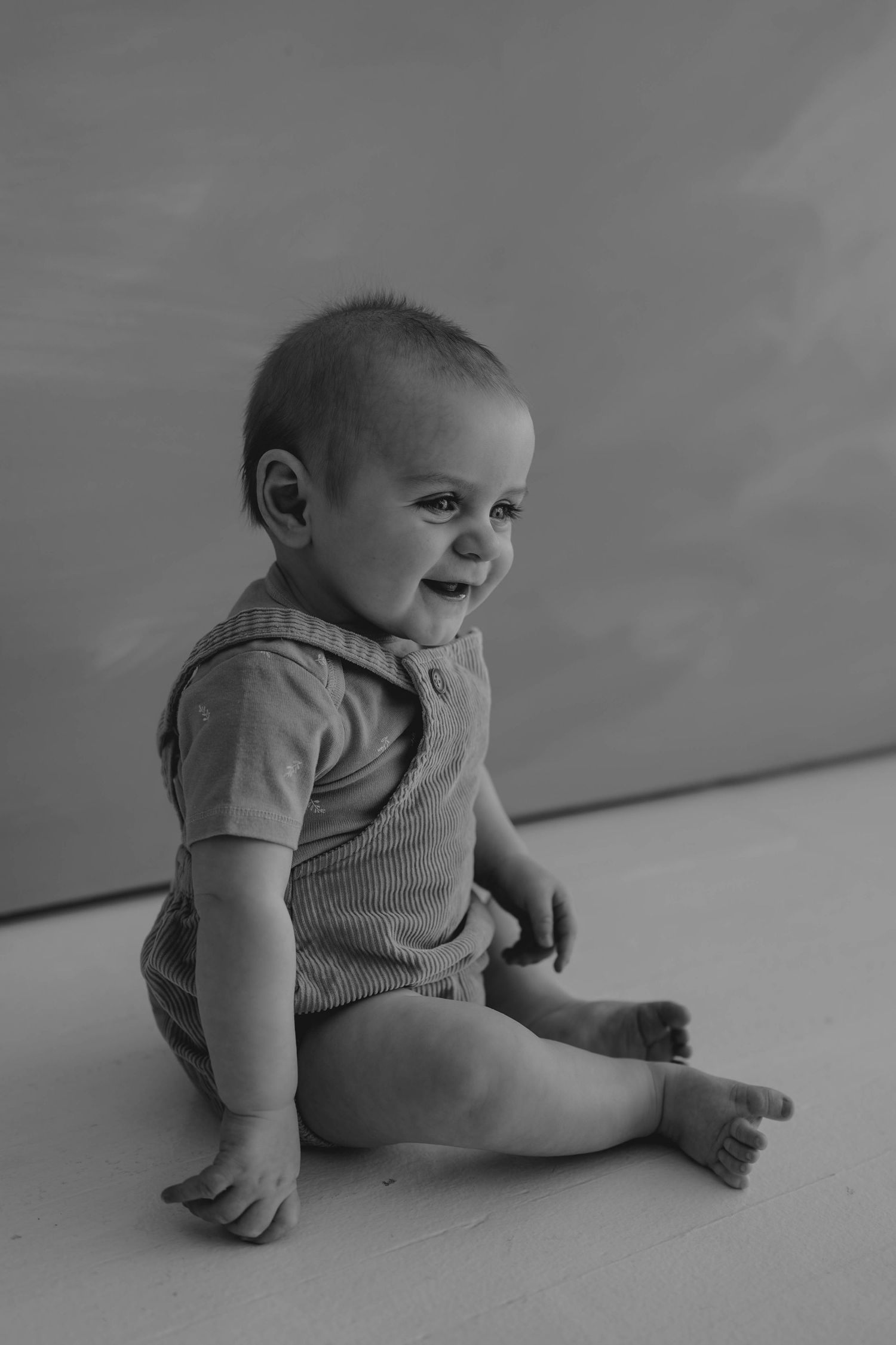 Black and white portrait of a happy smiling baby sitting on floor wearing a ribbed romper.