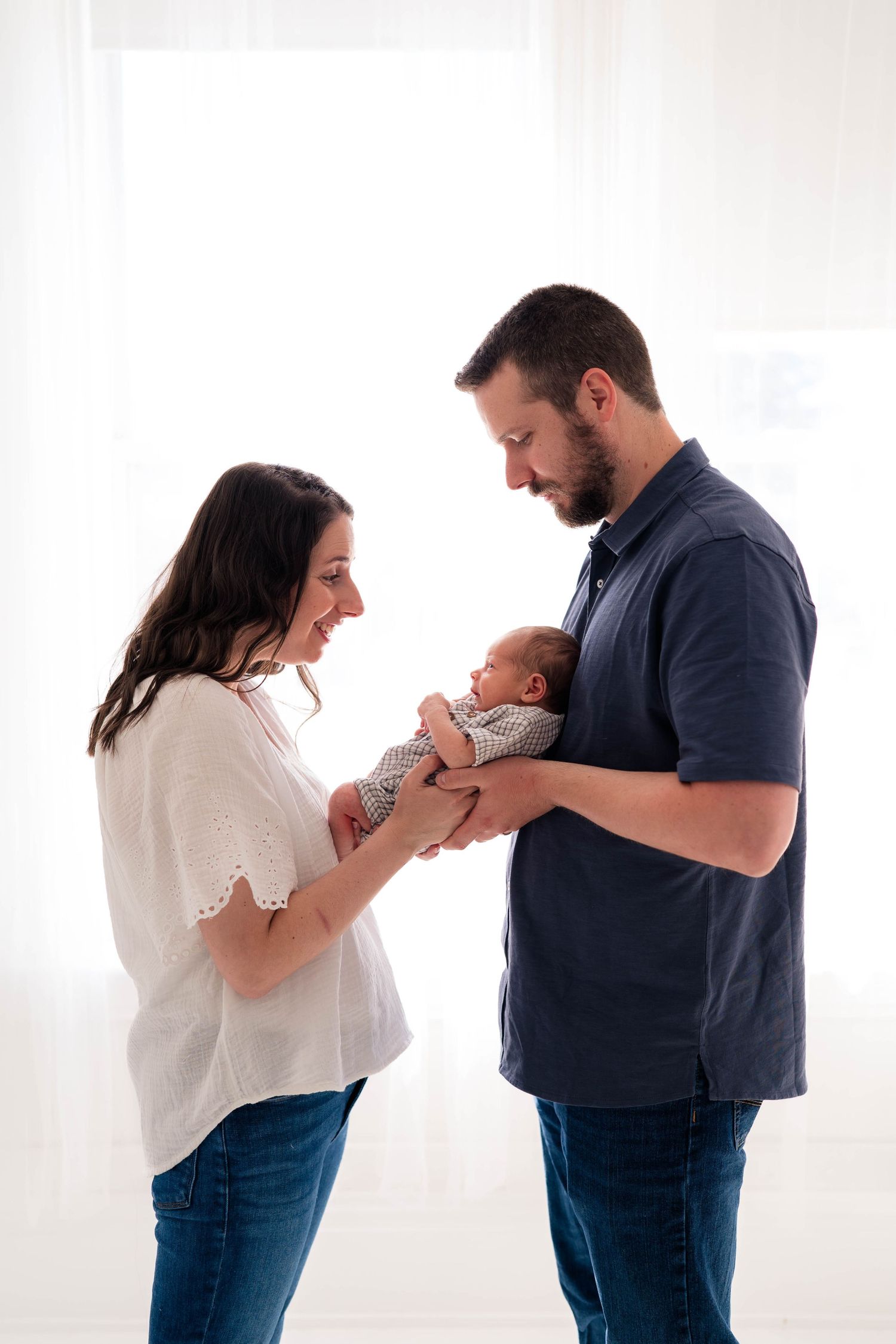 New parents standing together holding their sleeping newborn baby in front of a bright white window.